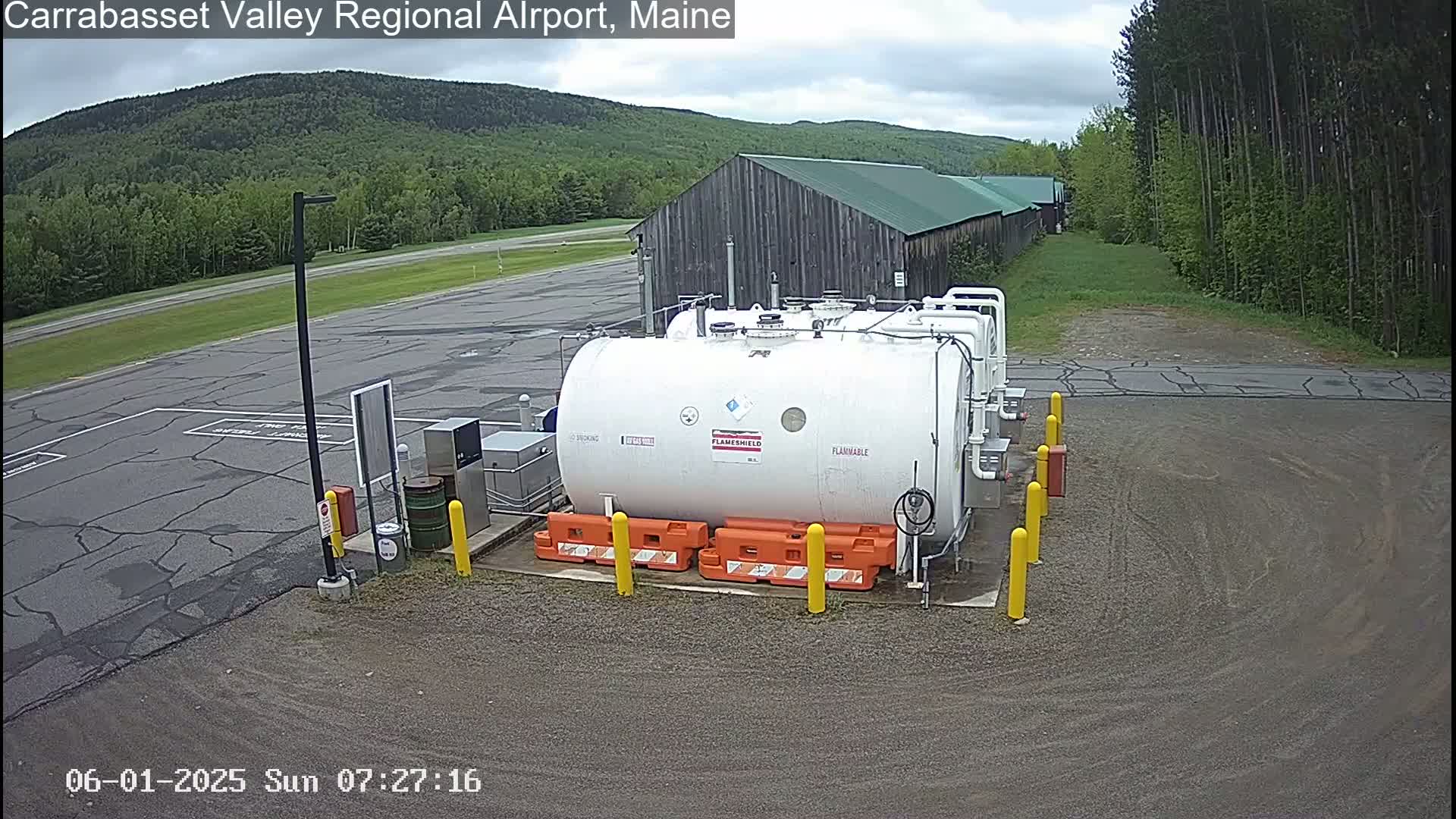 A large white fuel tank sits in a gravel area of an airport on an overcast day, with a wooden building and green trees in the background.