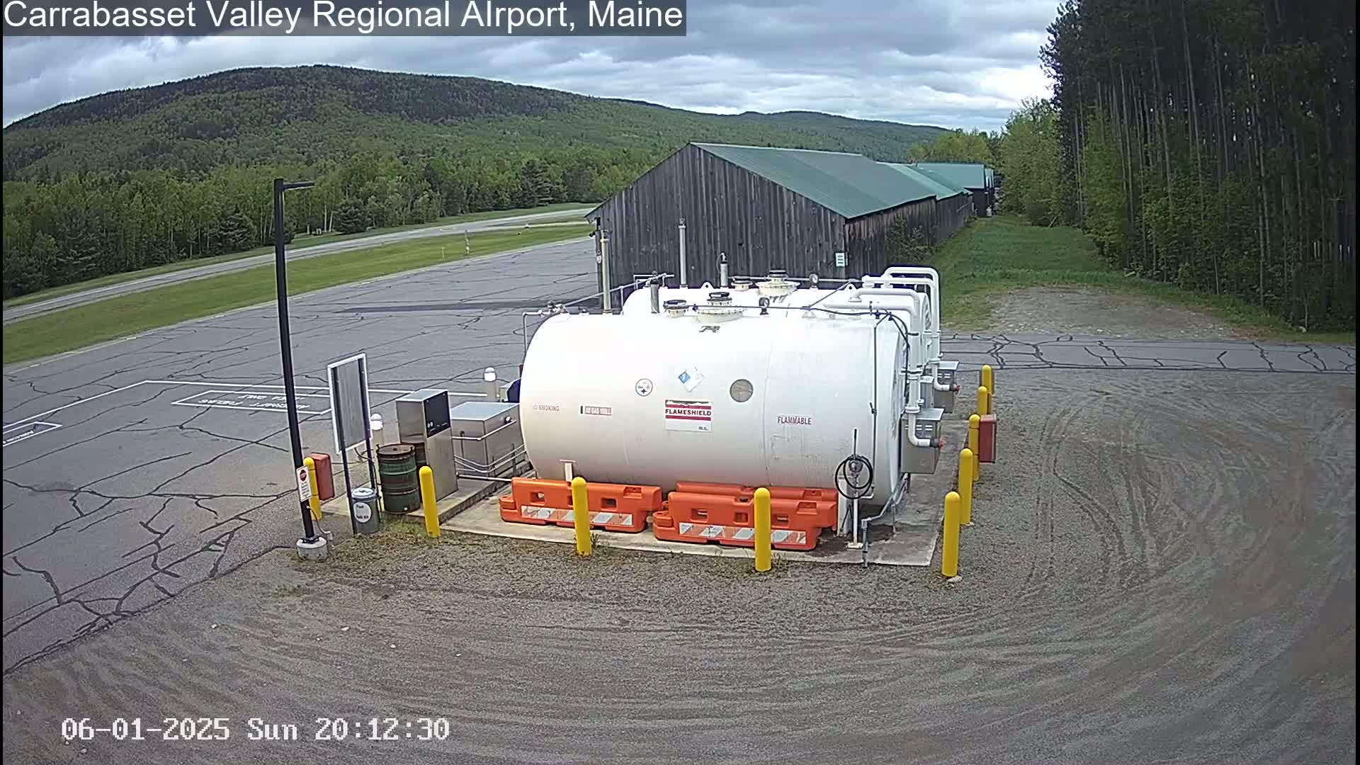 A large white fuel tank sits in a gravel area of an airport, under a partly cloudy sky, with a wood building and green trees in the background.