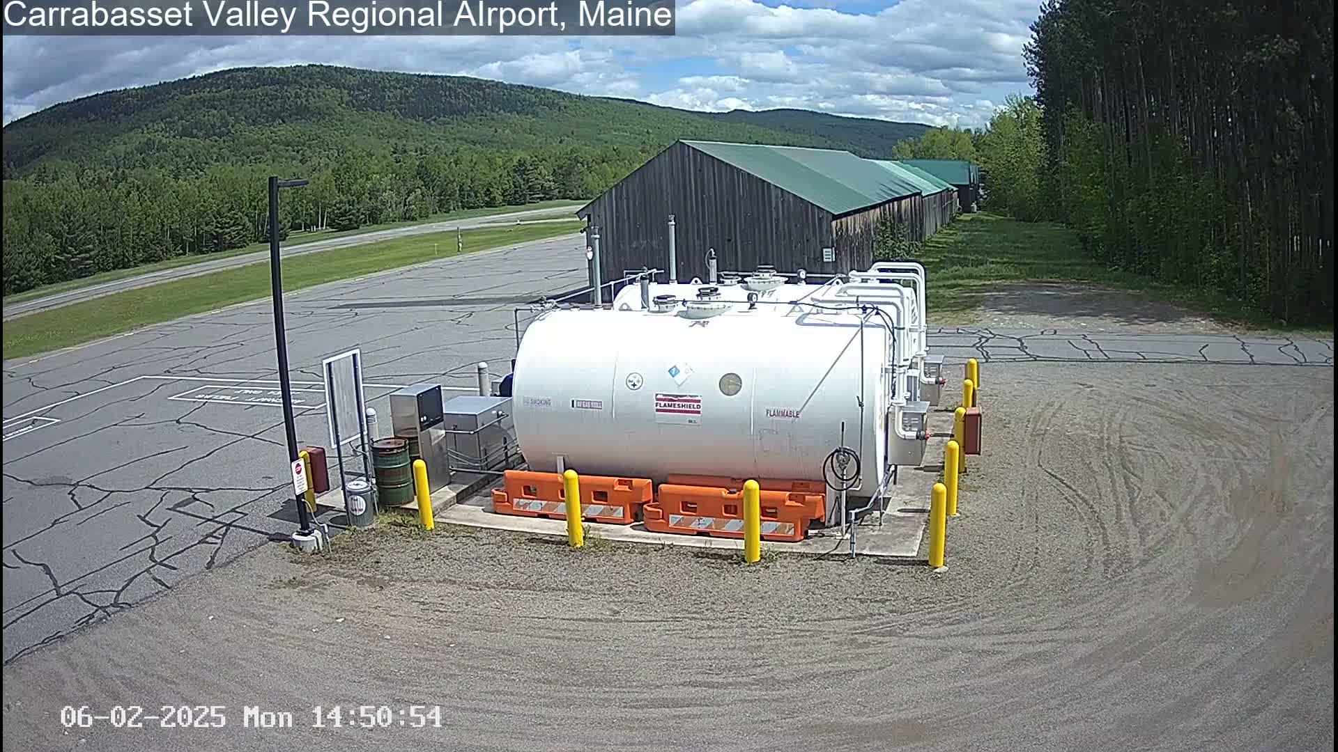 A large white fuel tank sits in a paved area of an airport, with a partly cloudy sky and green, tree-covered hills in the background.