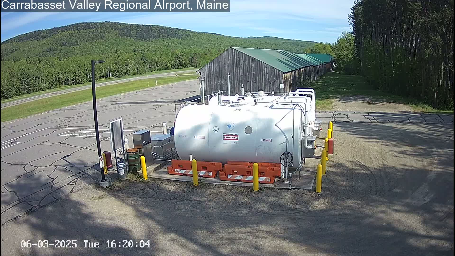 A large white fuel tank sits in a paved area of an airport, with a barn and forested hills visible in the background under a sunny sky.