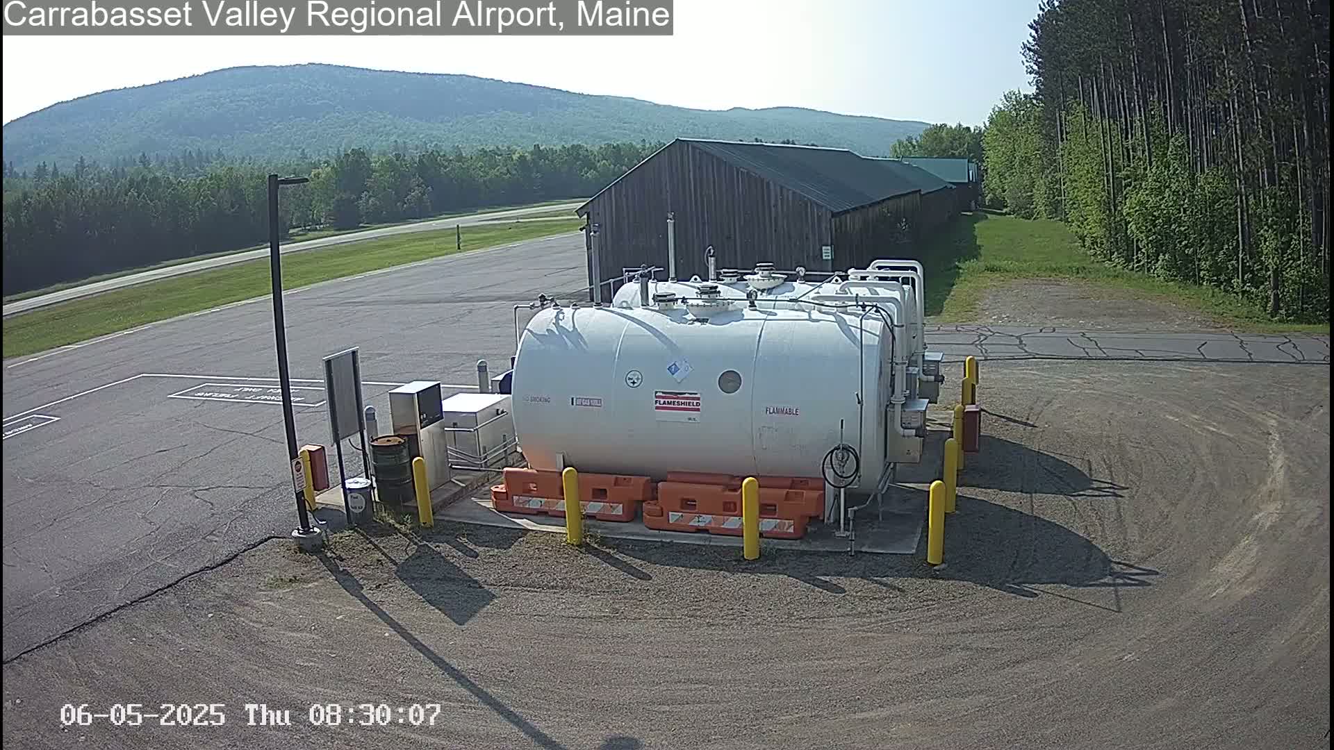 A large white cylindrical fuel tank sits in a paved area of an airport on a sunny day, with a brown building and green trees in the background.