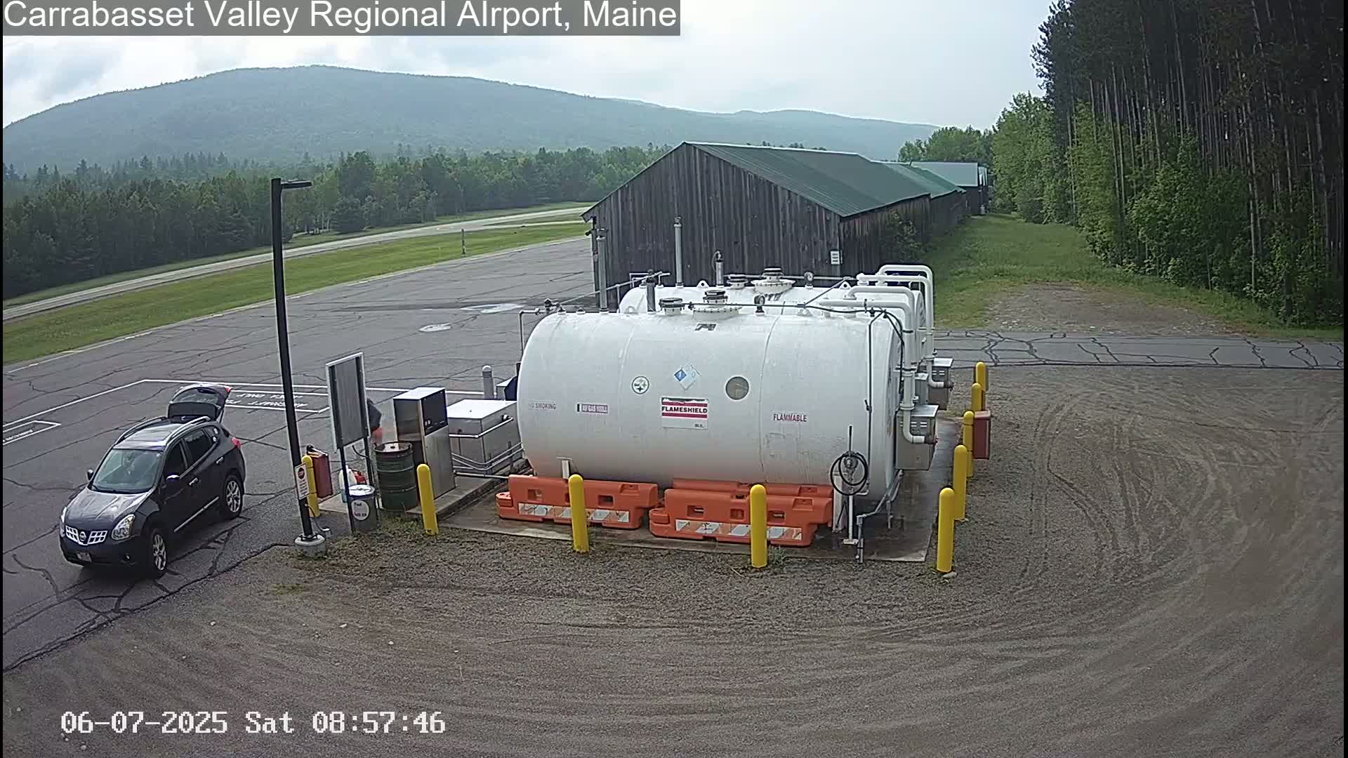 A dark-colored SUV is parked near a large white fuel tank in an airport parking lot on an overcast day, with a wooded area and distant mountains in the background.