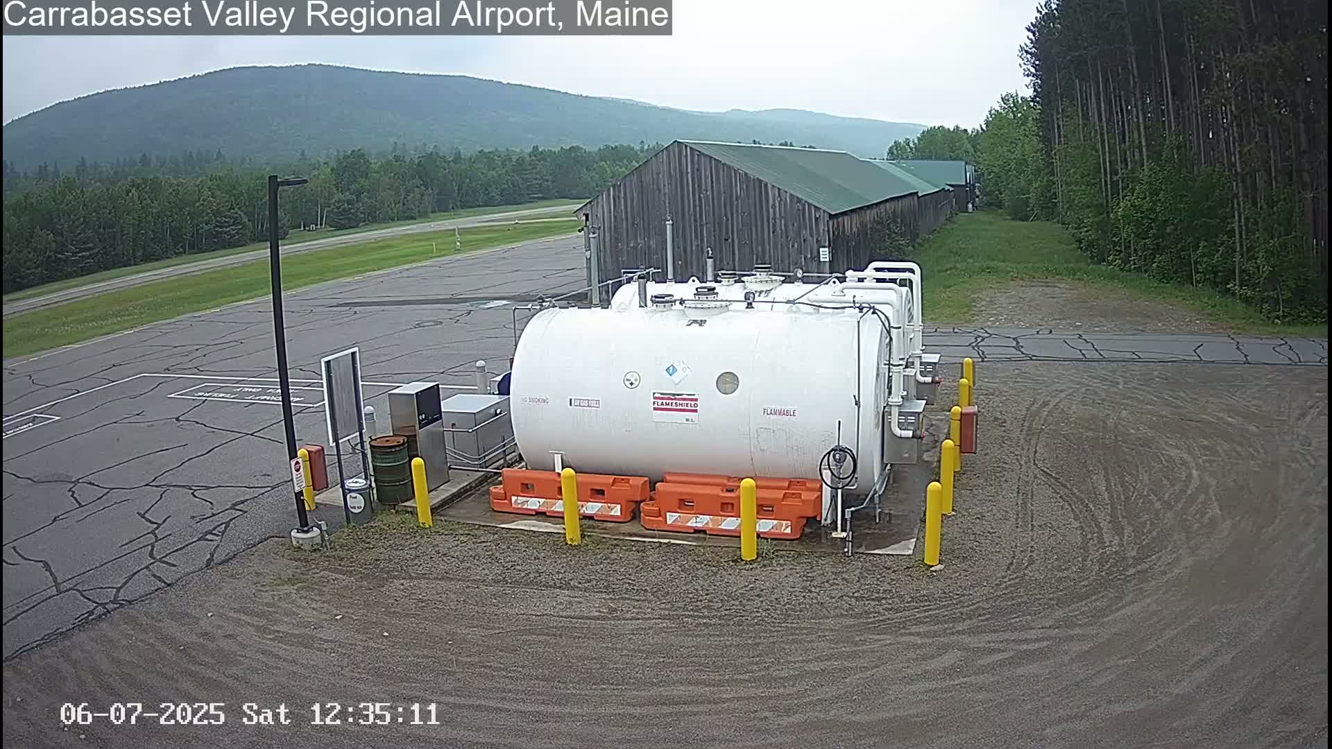 A large white cylindrical tank labeled "flammable" sits in a gravel area of an airport, under an overcast sky, with a wood building and green trees in the background.