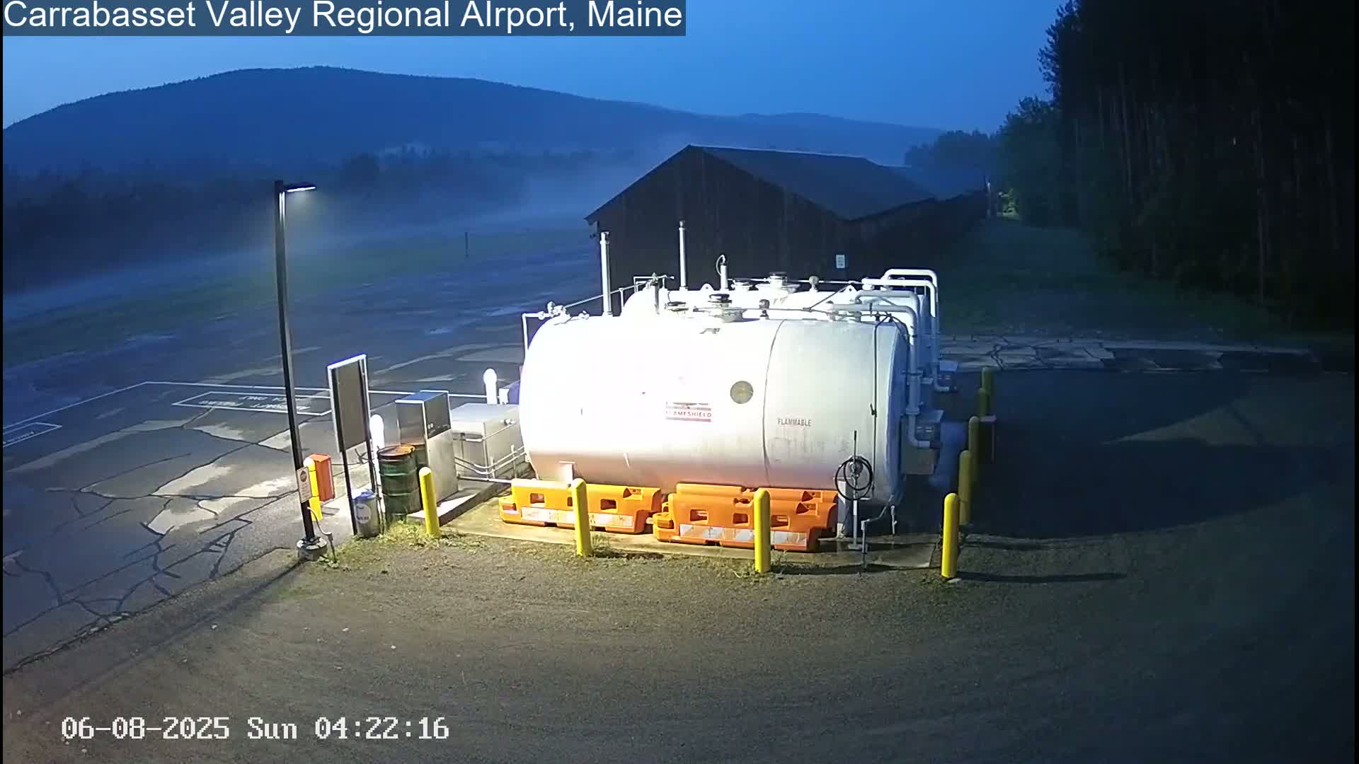 A large white cylindrical tank sits in a foggy parking lot at an airport, with a dark building and distant mountains visible in the low-light conditions.
