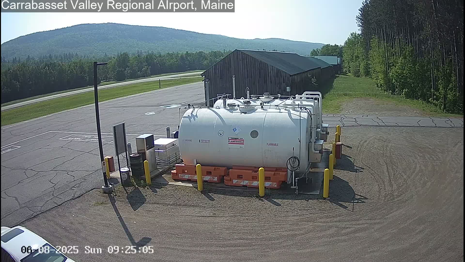 A large white fuel tank sits in a paved area near a long, dark-colored building, under a sunny sky with a mountain in the background.
