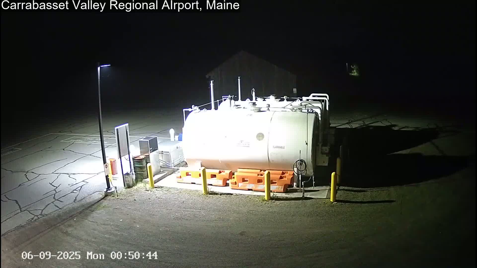 A large, white, flammable liquid storage tank sits in a dark, asphalt lot illuminated by a single lamppost at night.
