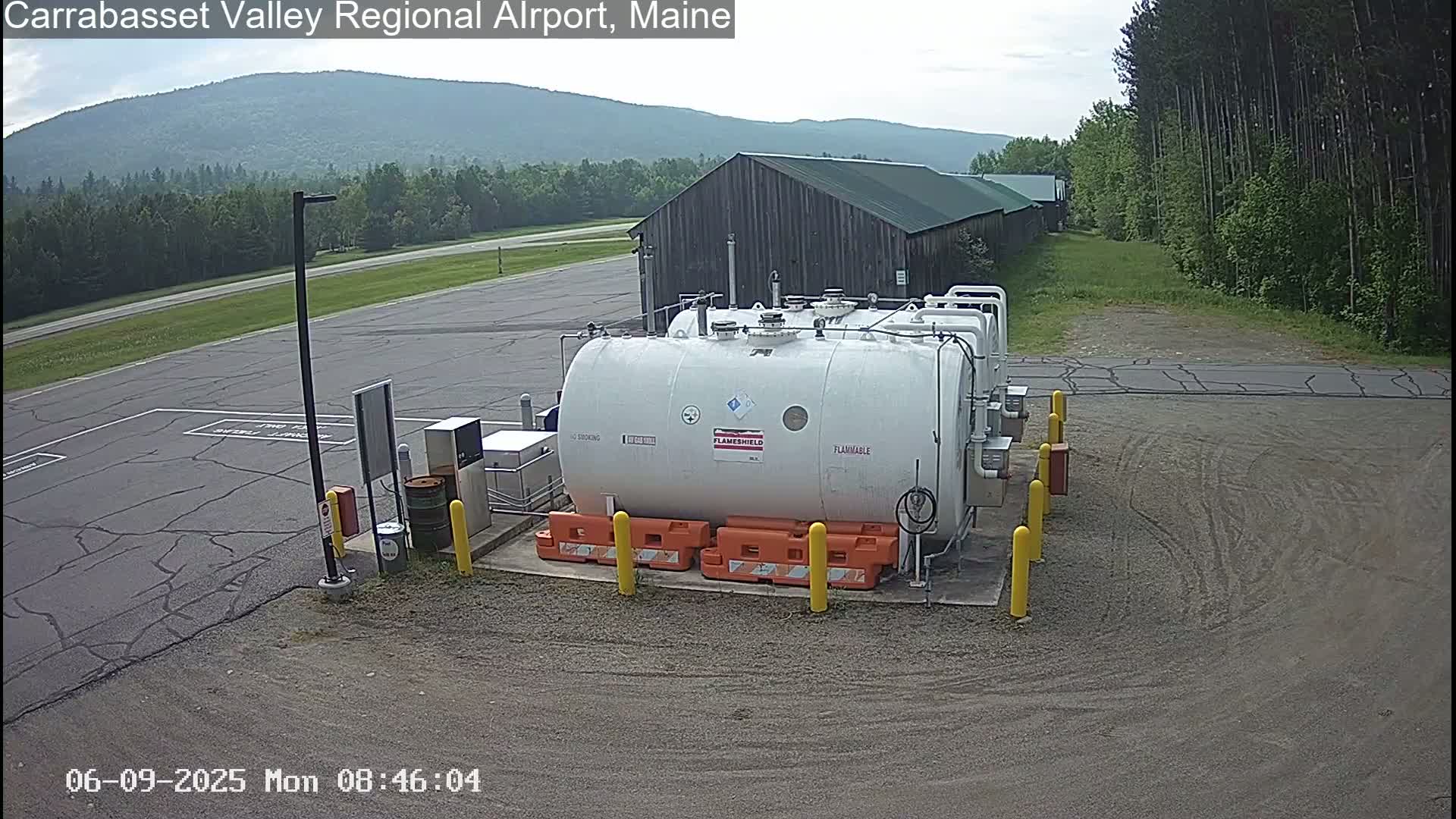 A large white fuel tank sits in a paved area near a wood building, under a mostly clear sky, with green hills and trees in the background.