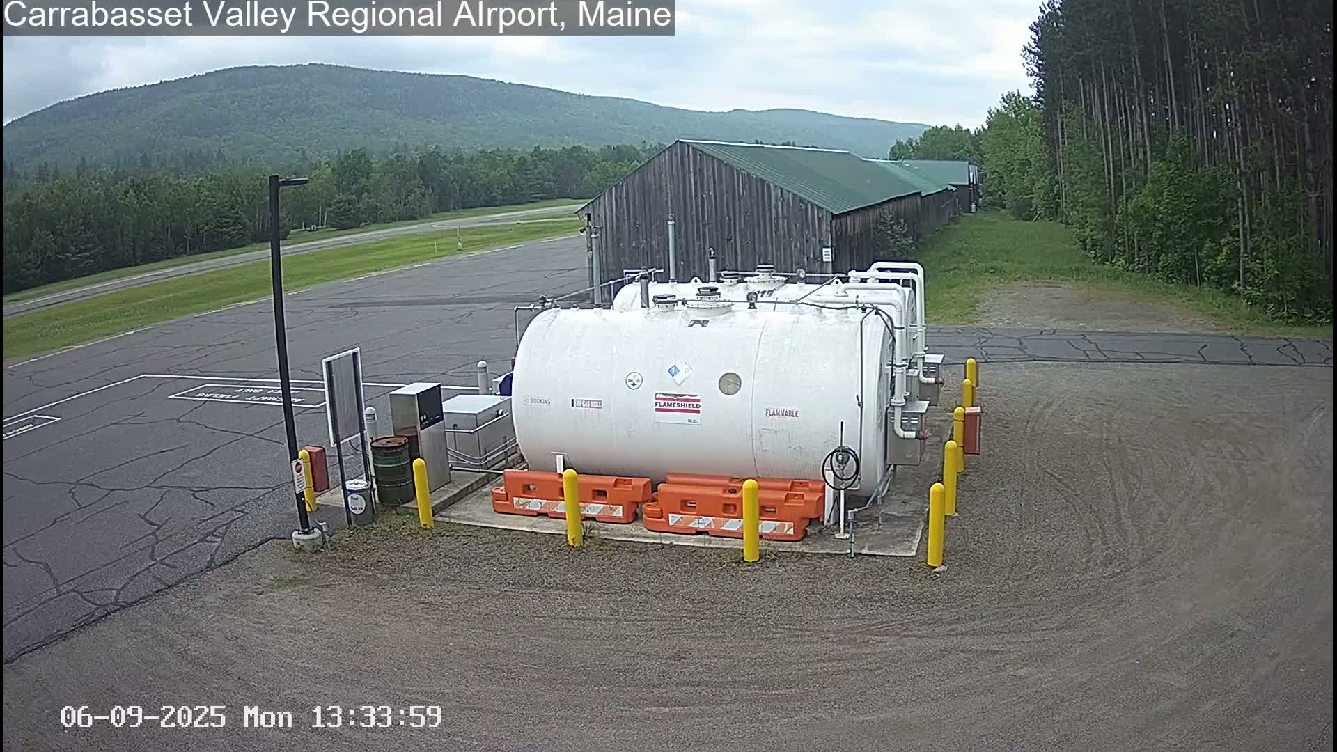 A large white flammable liquid storage tank sits in a paved area of an airport, with a grey building and green trees in the background under a partly cloudy sky.