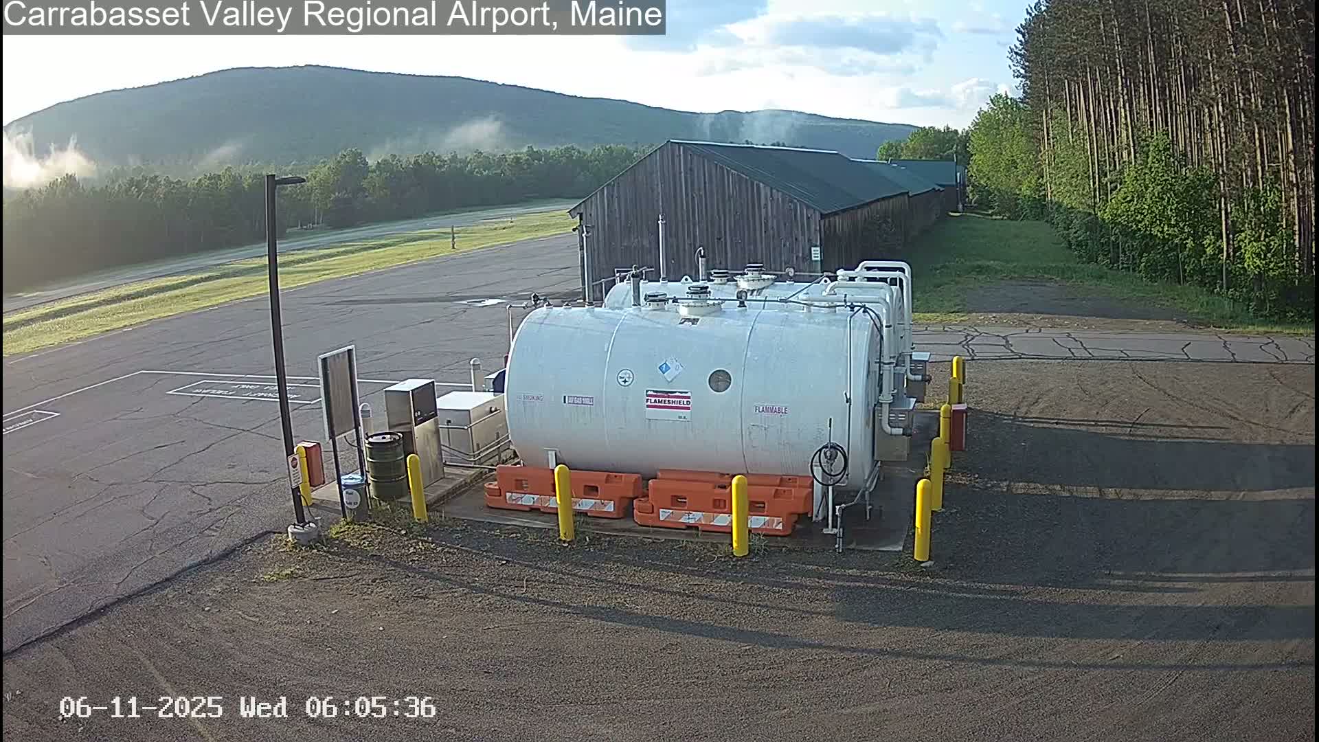 A large white fuel tank sits in a paved area near a wood building and a line of trees, with a light morning mist visible in the background.