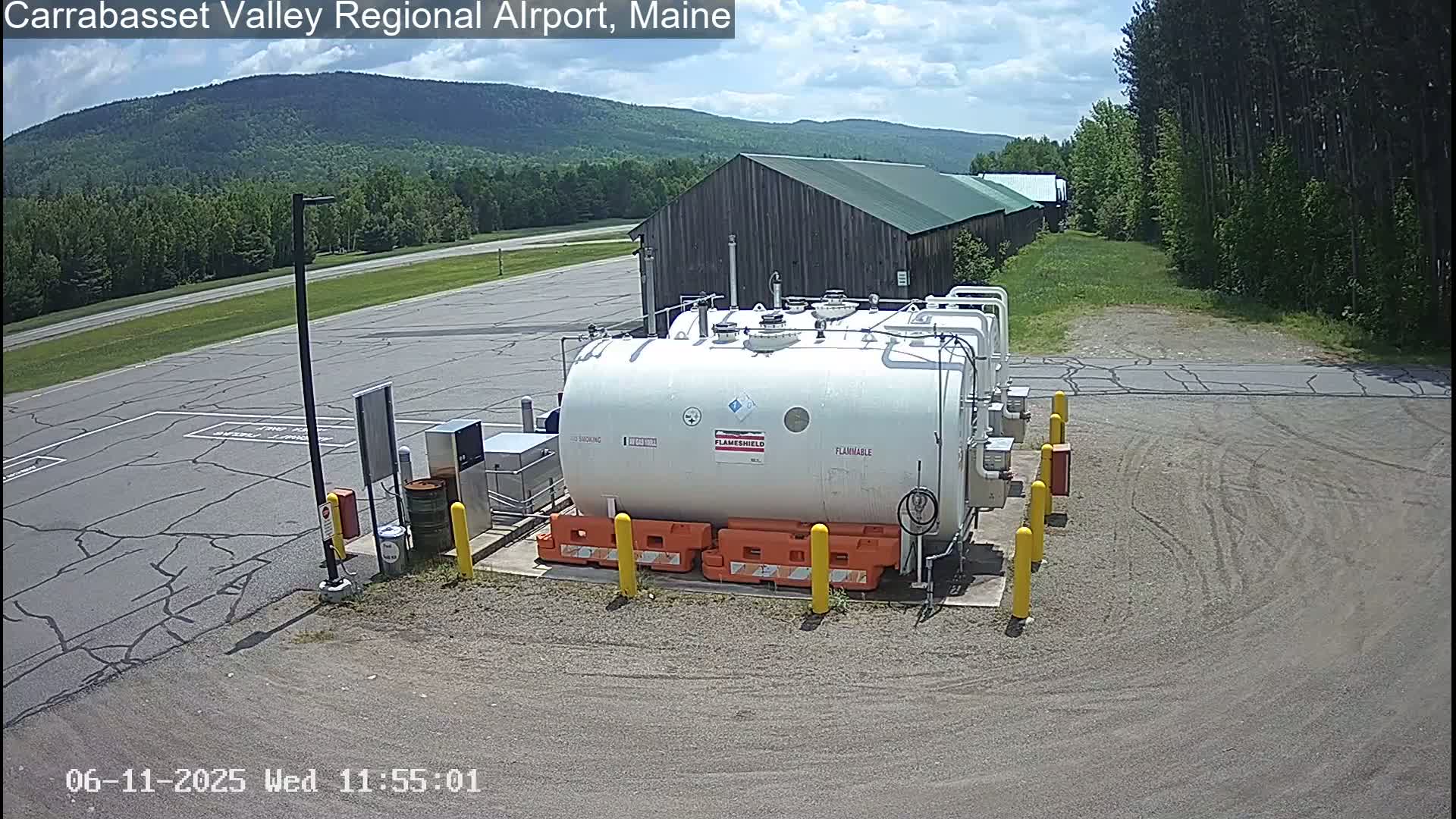 A large white cylindrical tank sits in a paved area next to a long, low building, under a partly cloudy sky, with green trees and distant mountains in the background.
