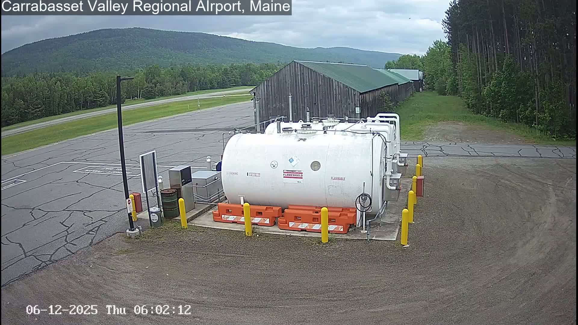 A large white cylindrical tank labeled "flammable" sits in a gravel area of an airport, with a wood barn and green forested hills in the background under an overcast sky.