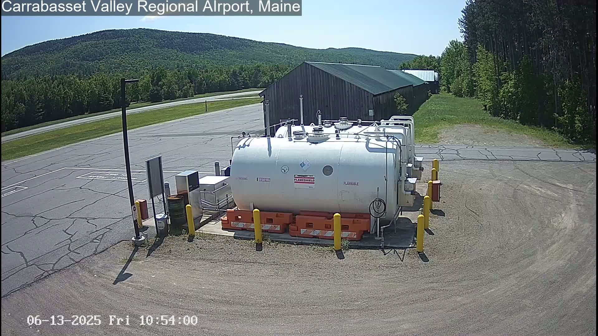 A large white fuel tank sits in a paved area near a wood building, under a sunny sky, with green forested hills in the background.