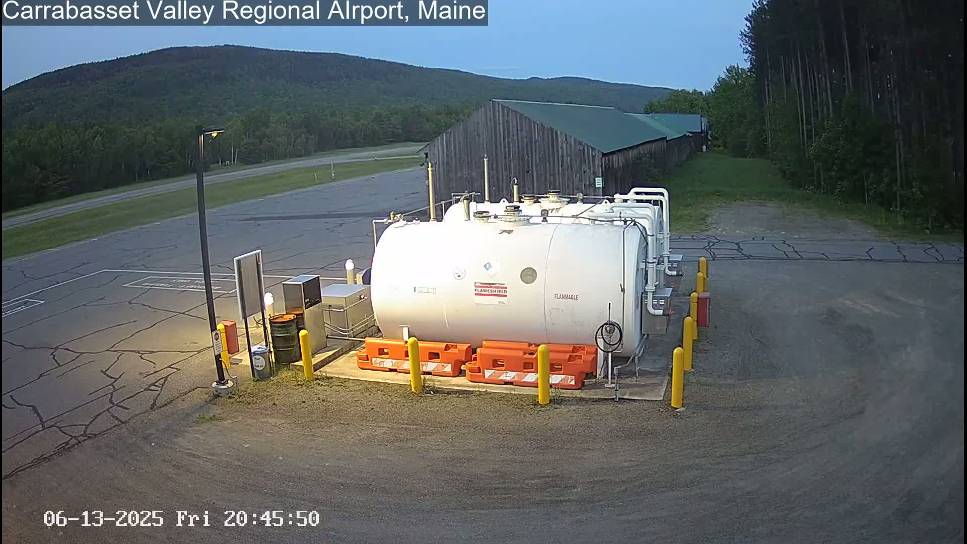 A large white fuel tank sits in a paved area near a wood building, under a sunny sky, with green forested hills in the background.