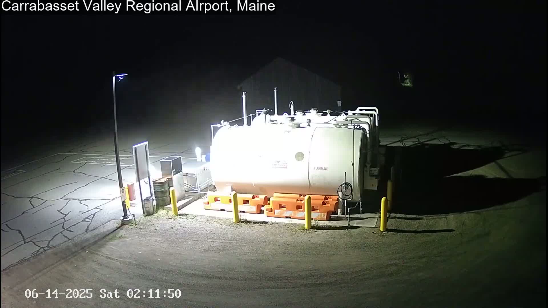 A large white fuel tank sits in a dark, nighttime airport lot illuminated by a single lamppost.