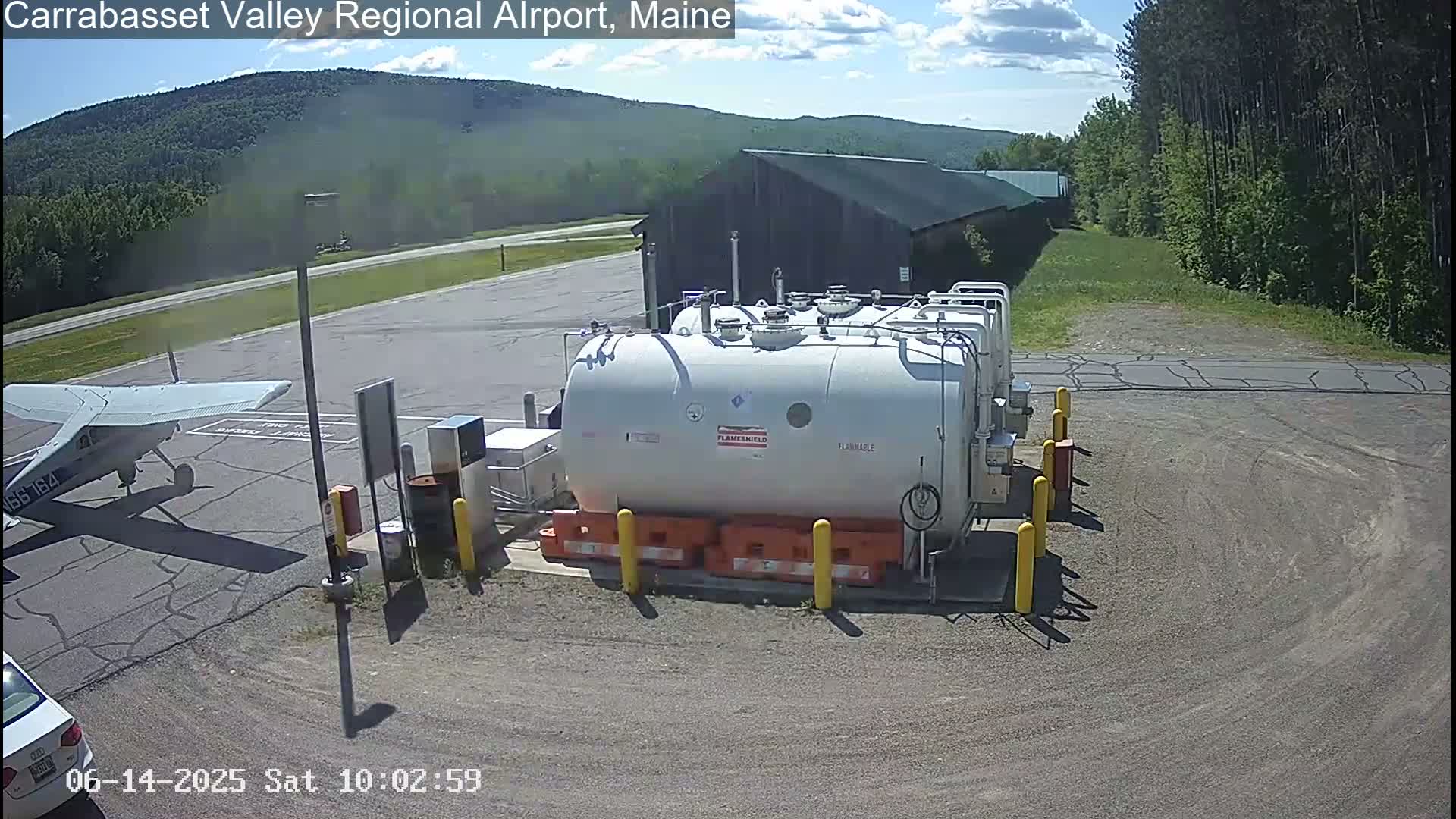 A small airplane is parked near a fuel depot at an airport on a sunny day, with green hills and trees in the background.