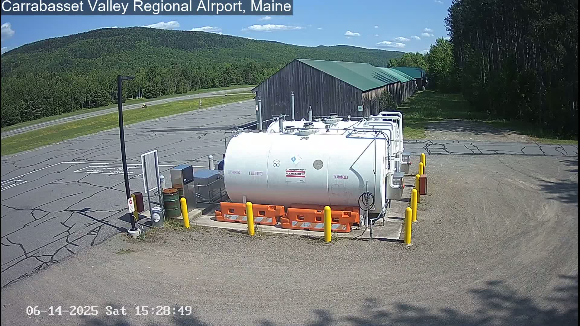 A small airplane is parked near a fuel depot at an airport on a sunny day, with green hills and trees in the background.
