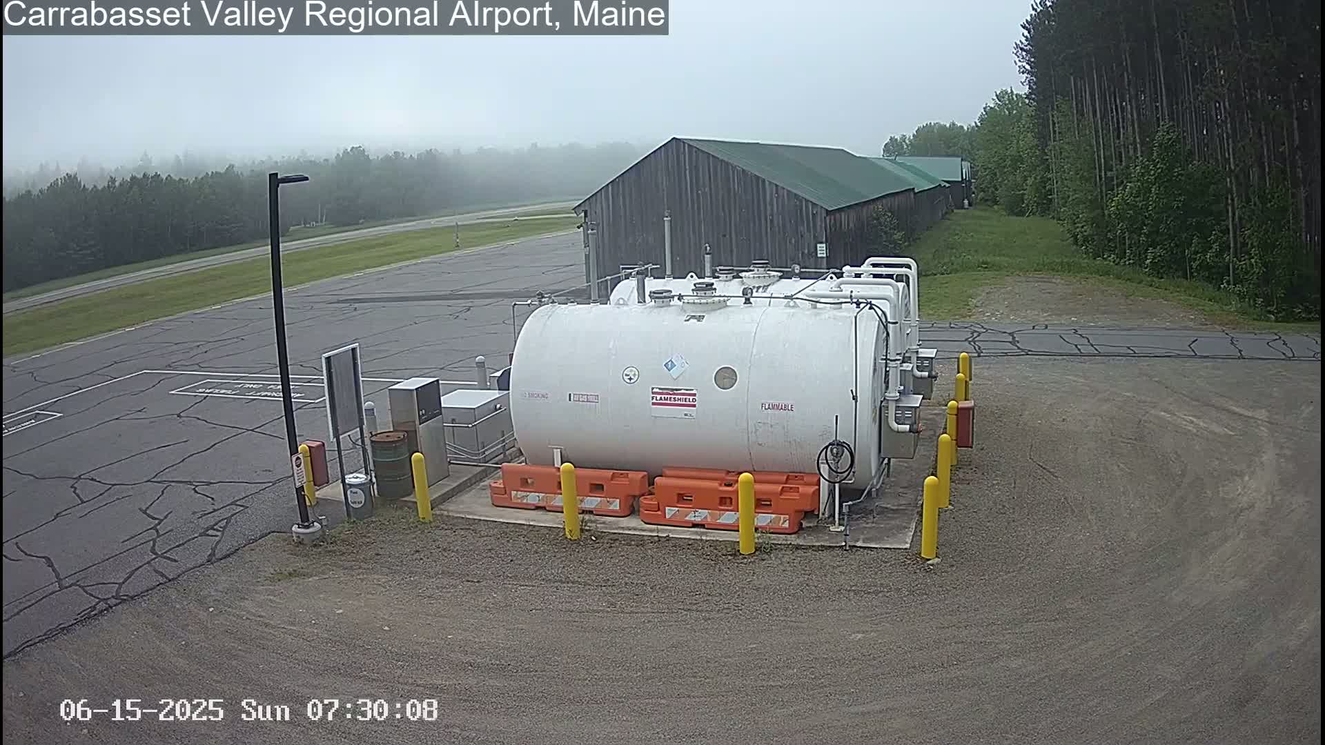 A large white fuel tank sits in a foggy airport parking lot next to a wood-sided building.