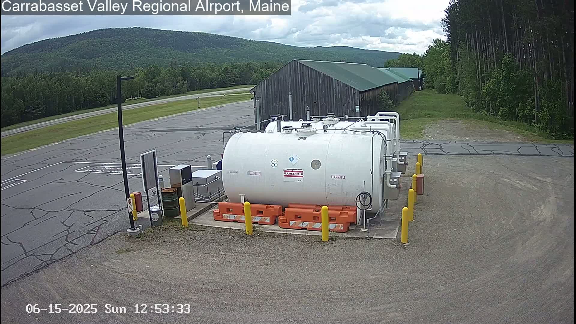 A large white fuel tank sits in a paved area of an airport, with a barn and a partly cloudy sky visible in the background.