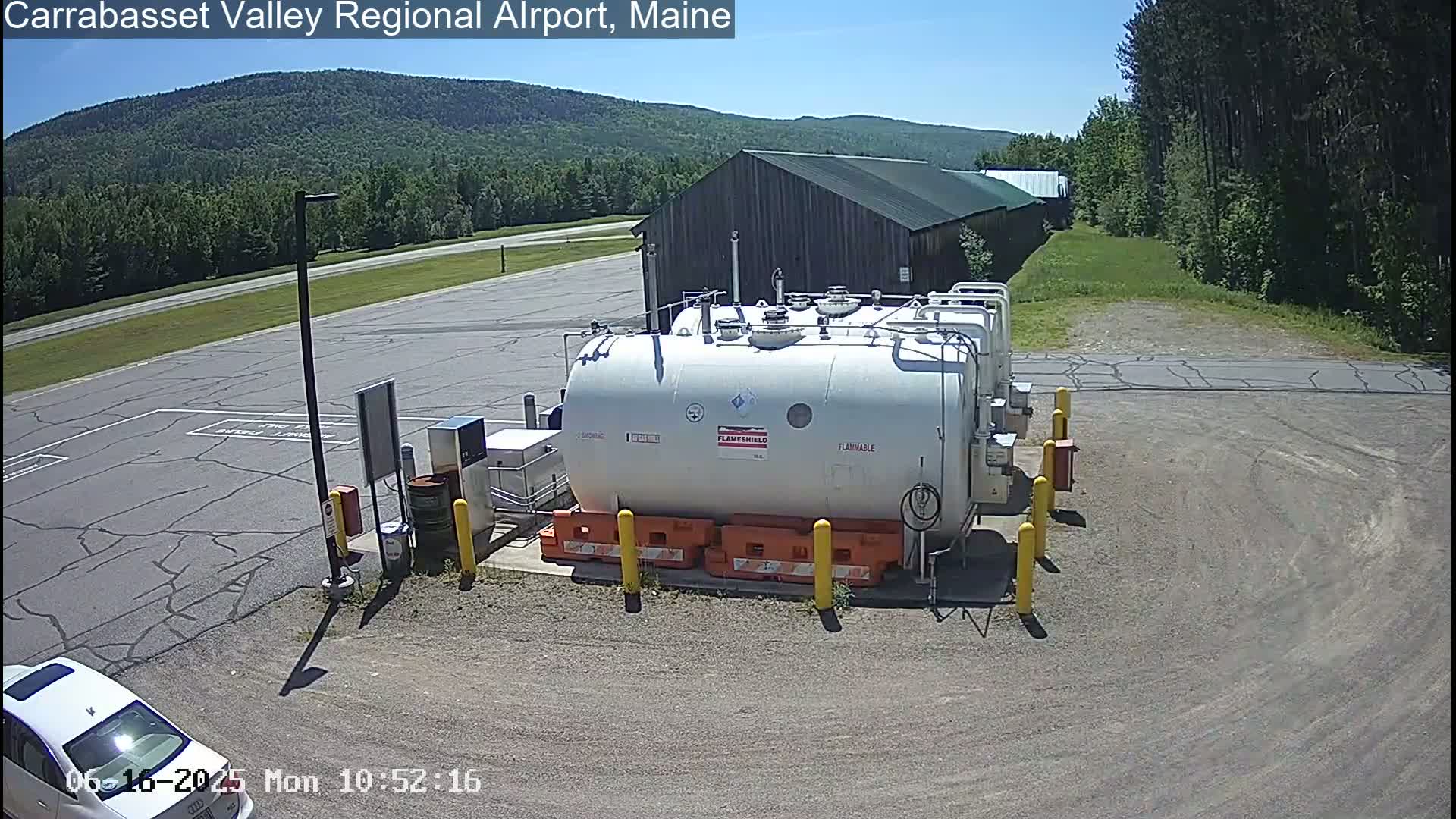 A large white flammable liquid storage tank sits in a paved area of an airport on a sunny day, with a wood-sided building and green hills in the background.