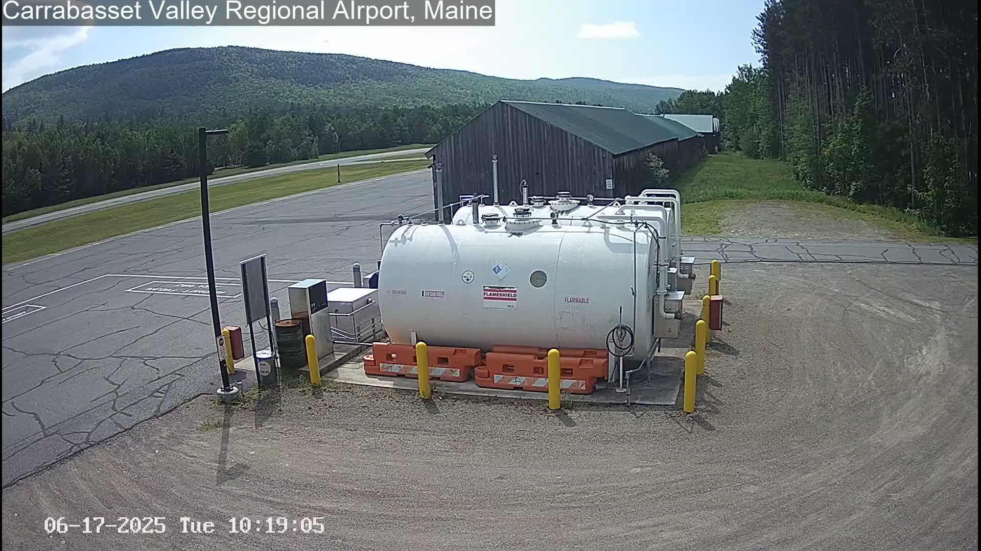 A large white fuel tank sits in a paved area next to a long, low wooden building, under a mostly sunny sky, with a background of green forested hills.