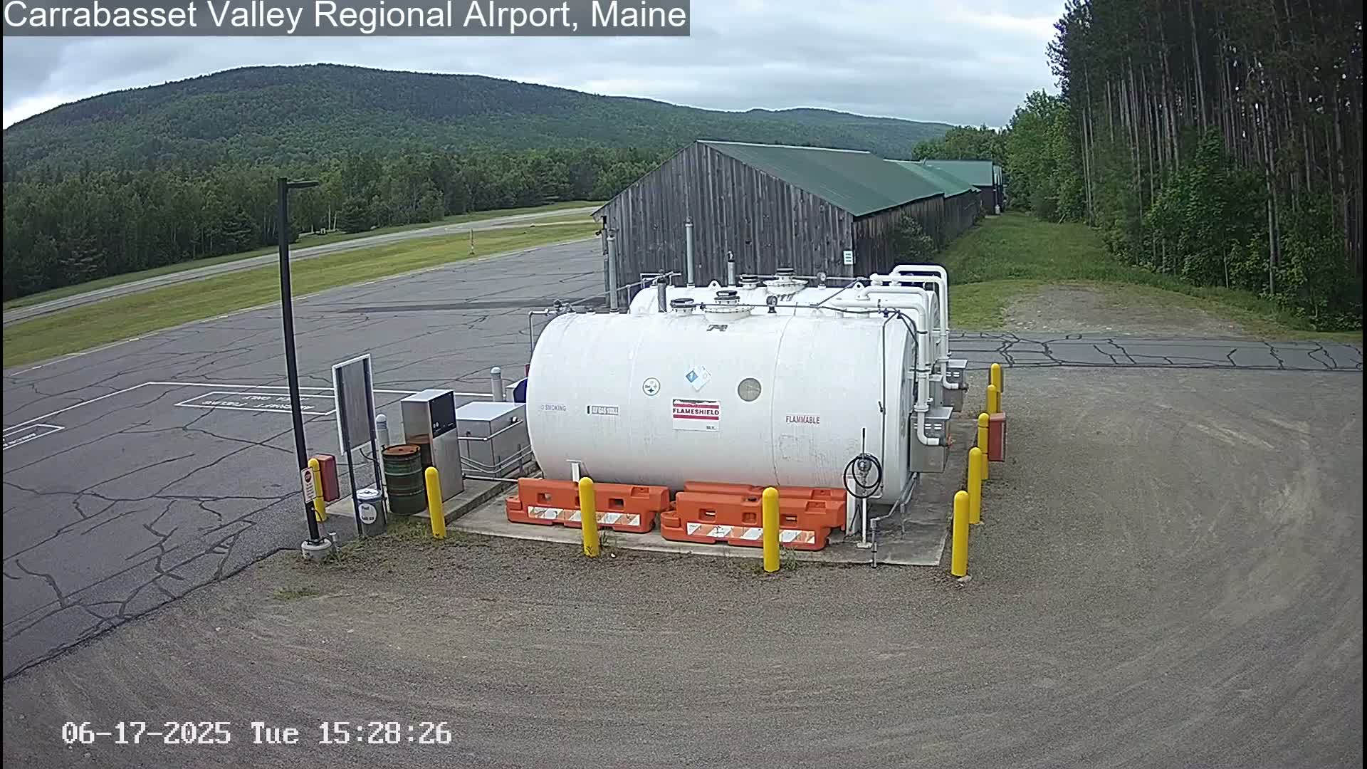 An outdoor scene on an overcast day shows a large white fuel tank surrounded by orange safety barriers in a parking lot next to a long wooden building, with a distant mountain range visible in the background.