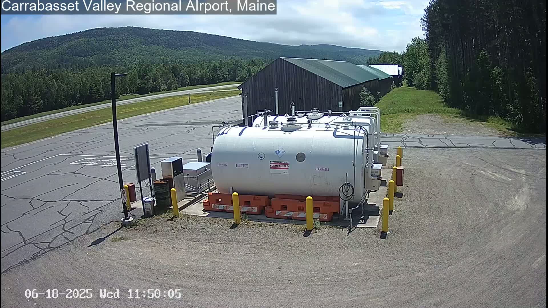 A large white fuel tank sits in a paved area of an airport, with a wood building and green mountains visible in the background under a mostly sunny sky.