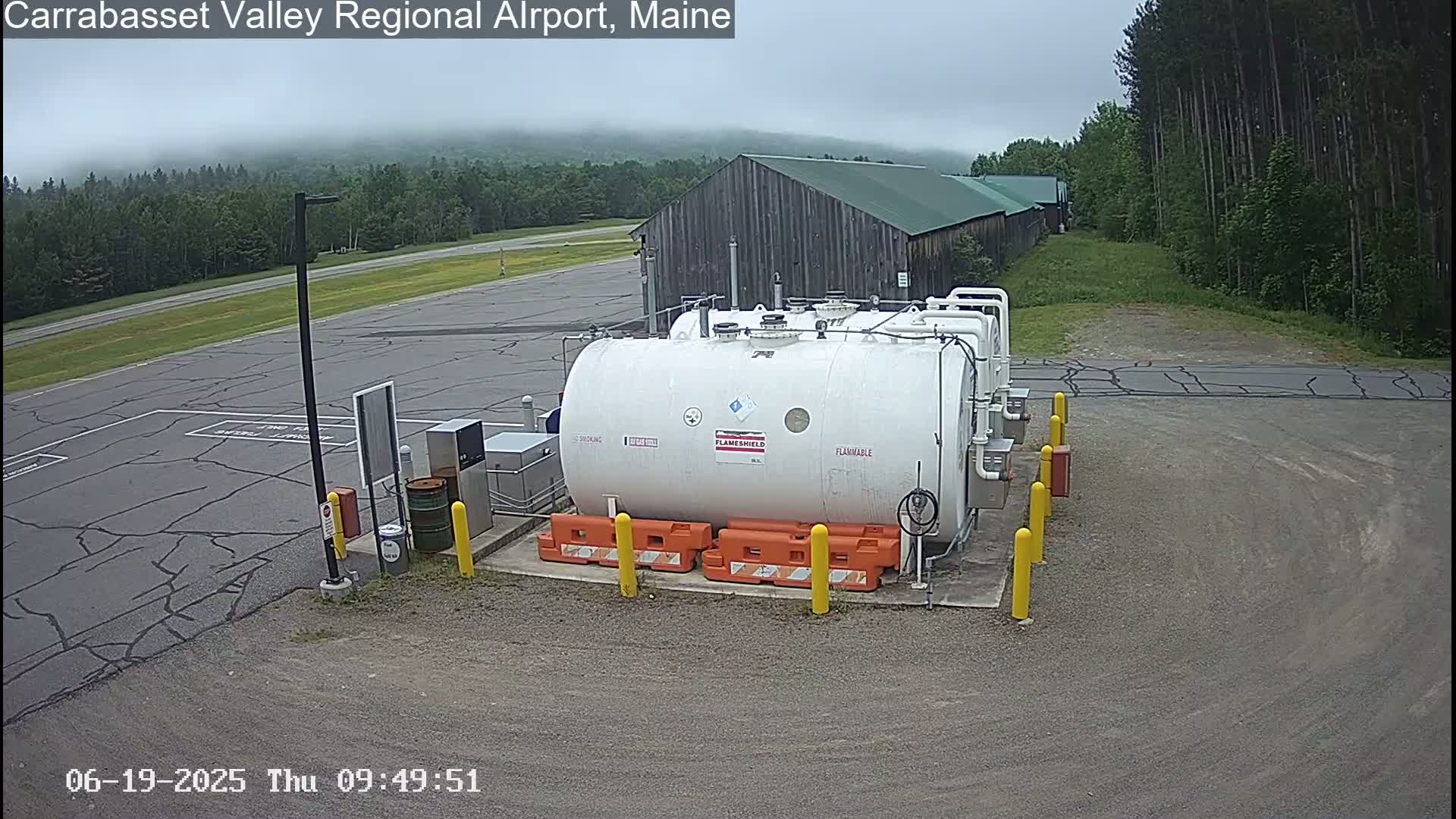 A large white flammable liquid storage tank sits in a mostly empty asphalt lot on an overcast day, near a long wooden building and a line of trees.