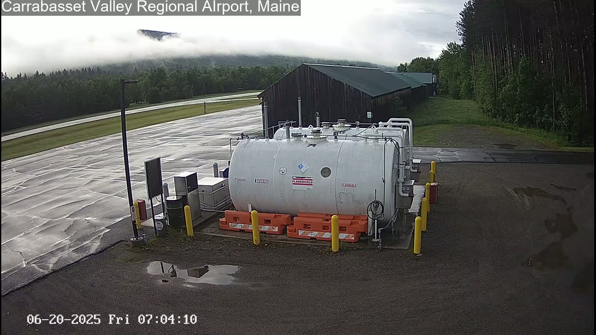 A large white cylindrical tank labeled "flammable" sits in a wet, paved area near a dark-colored building, with low clouds partially obscuring the distant hills.