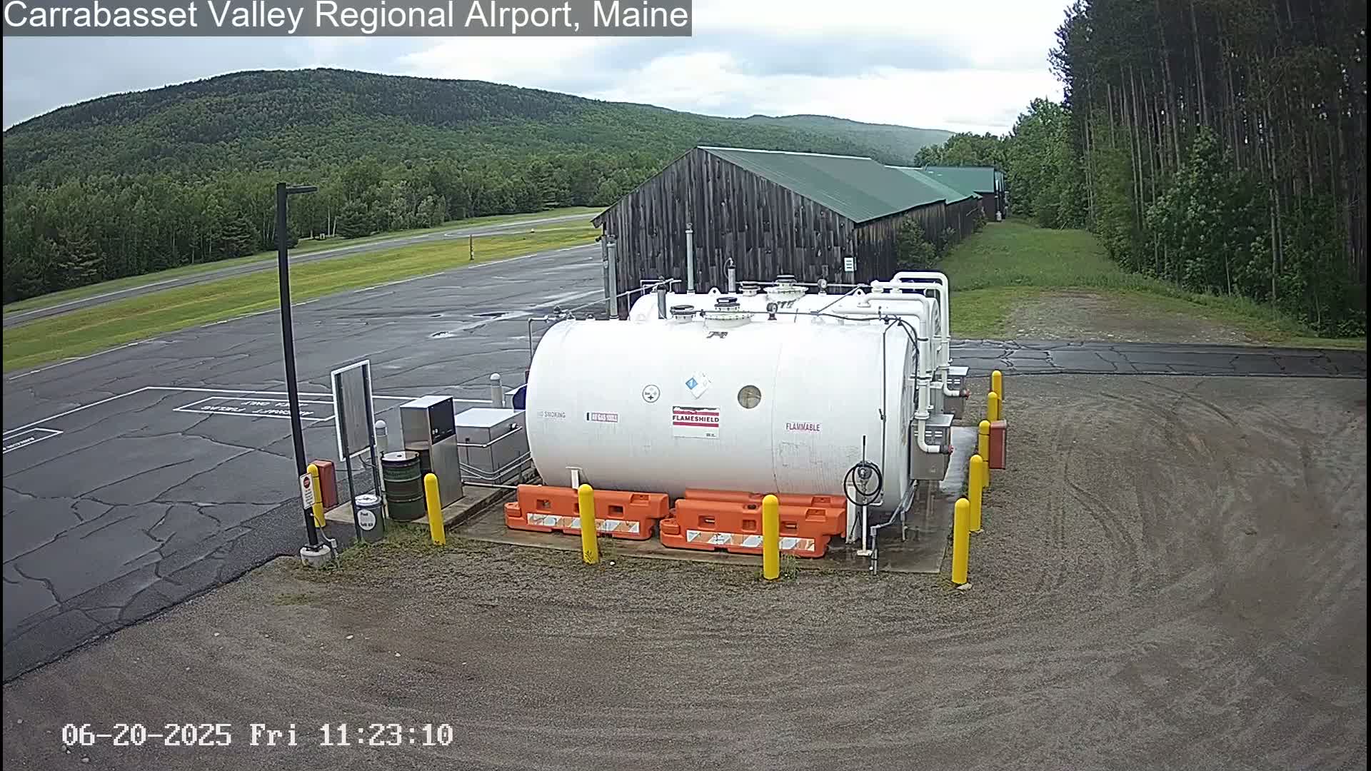 A large white fuel tank sits in a gravel area of an airport, with a dark-colored building and green, forested hills in the background under an overcast sky.