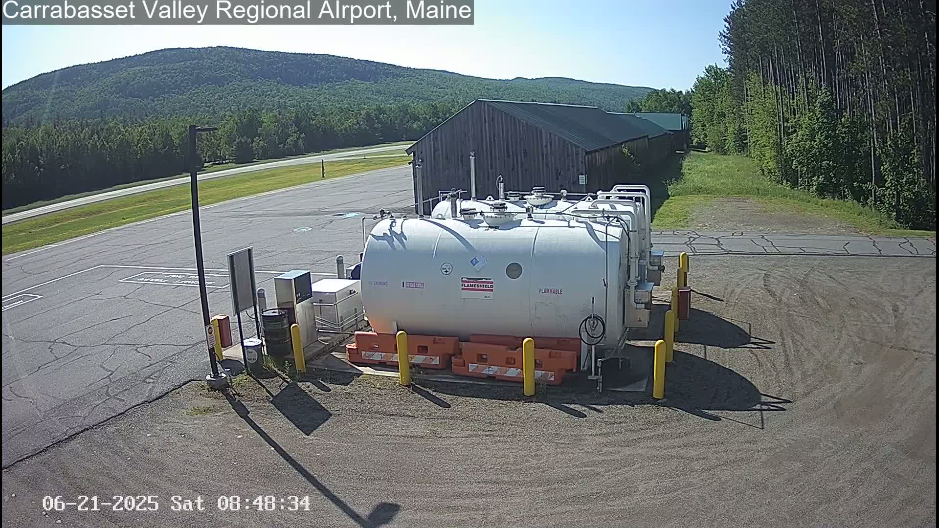 A large white fuel tank sits in a paved area near a brown building, under a clear sunny sky with green forested hills in the background.