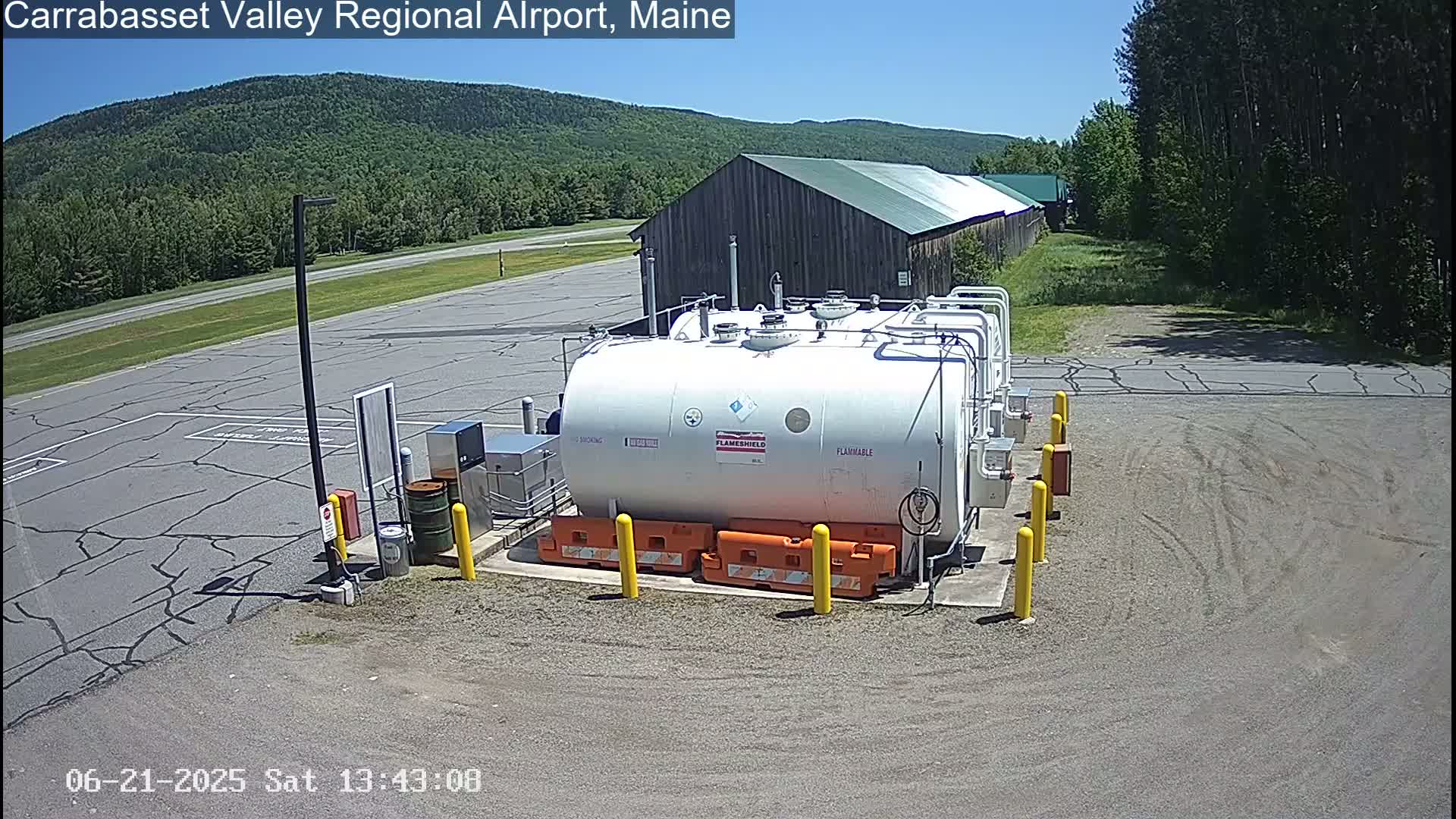 A large white fuel tank sits in a paved area of an airport on a sunny day, with a wooden building and green forested hills in the background.