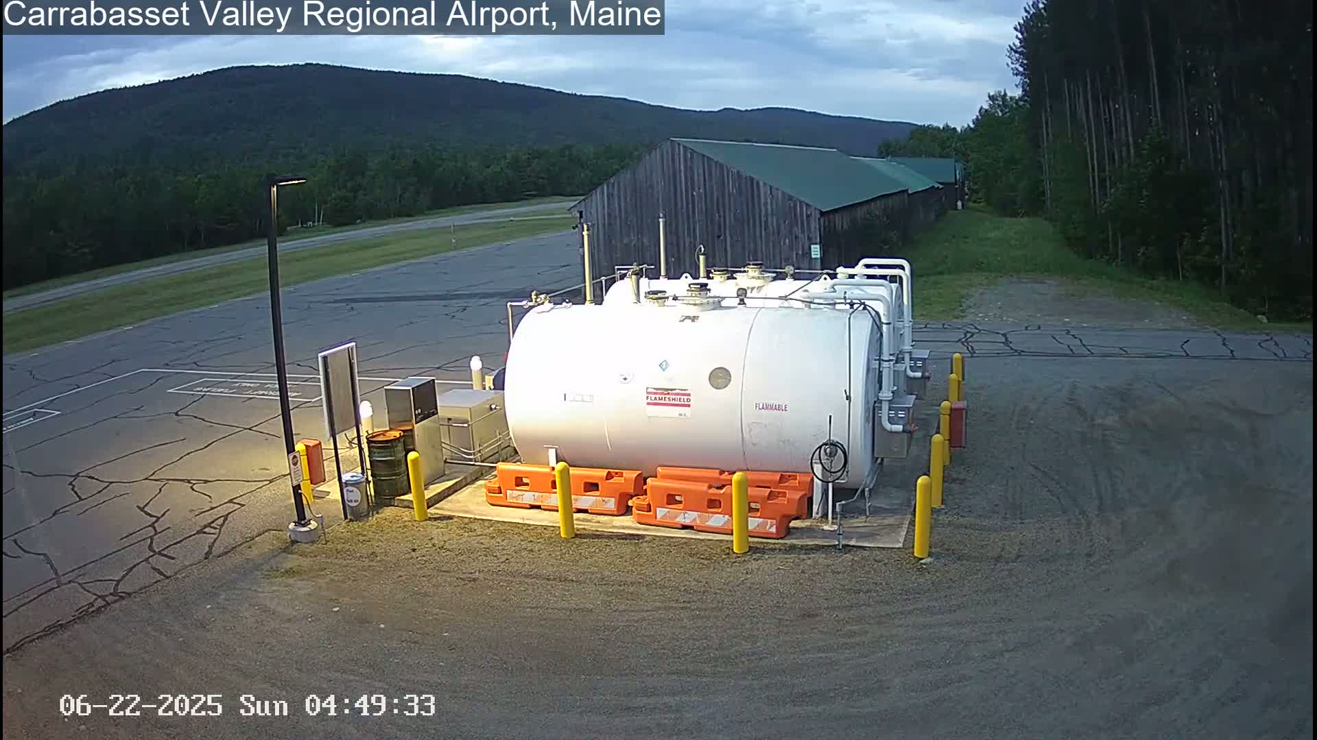 A large white cylindrical fuel tank sits in a paved area of an airport, with a long barn-like building and distant mountains visible under a partly cloudy sky.