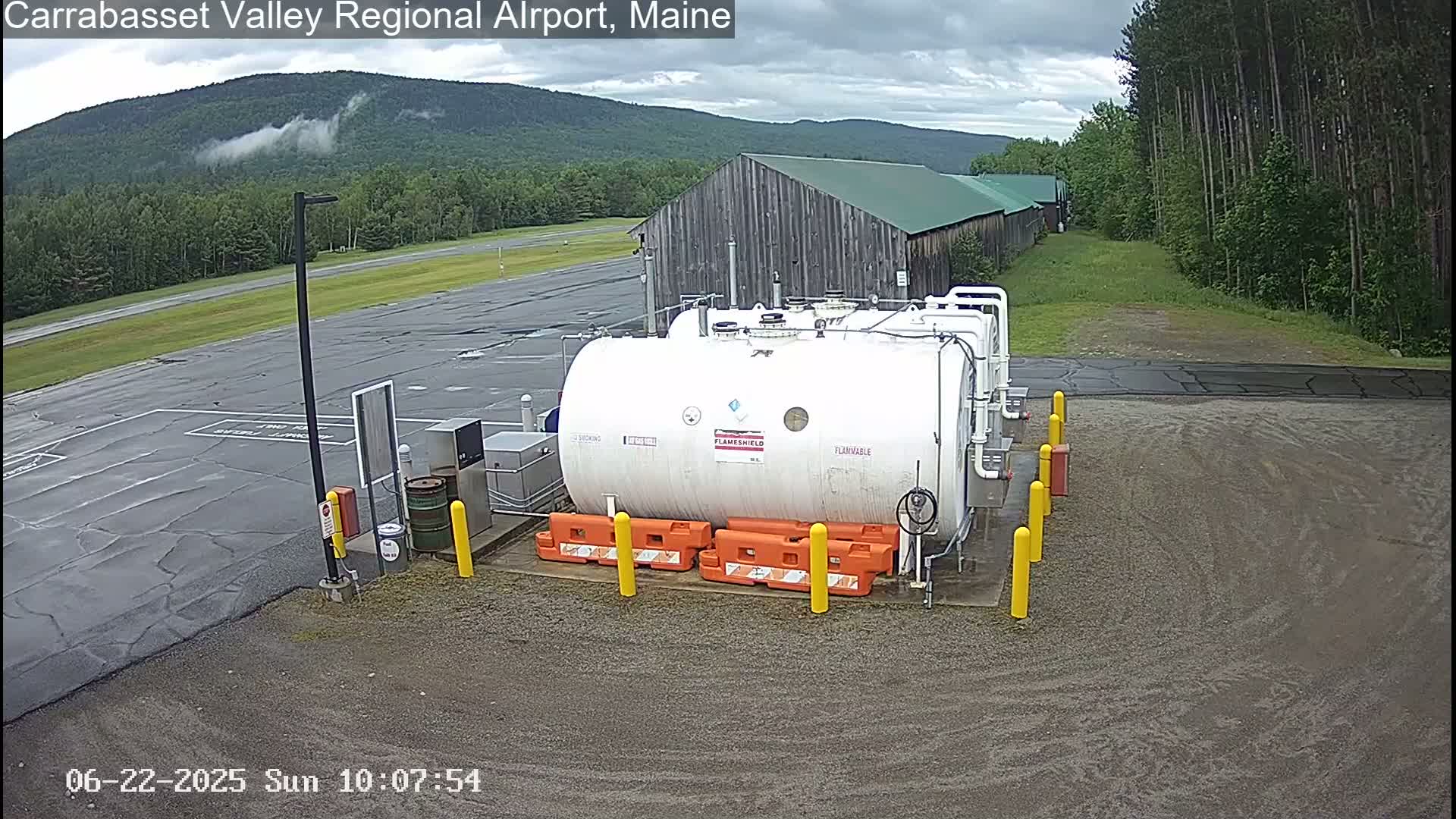 A large white fuel tank sits in a paved area of an airport, with a wood building and green, tree-covered mountains in the background under a cloudy sky.