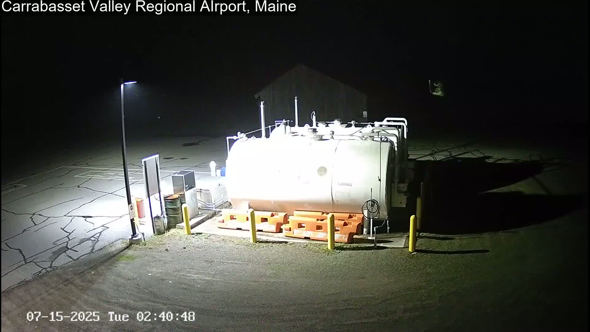 A large, cylindrical fuel tank sits in a dark, empty parking lot at night, illuminated by a single light pole.