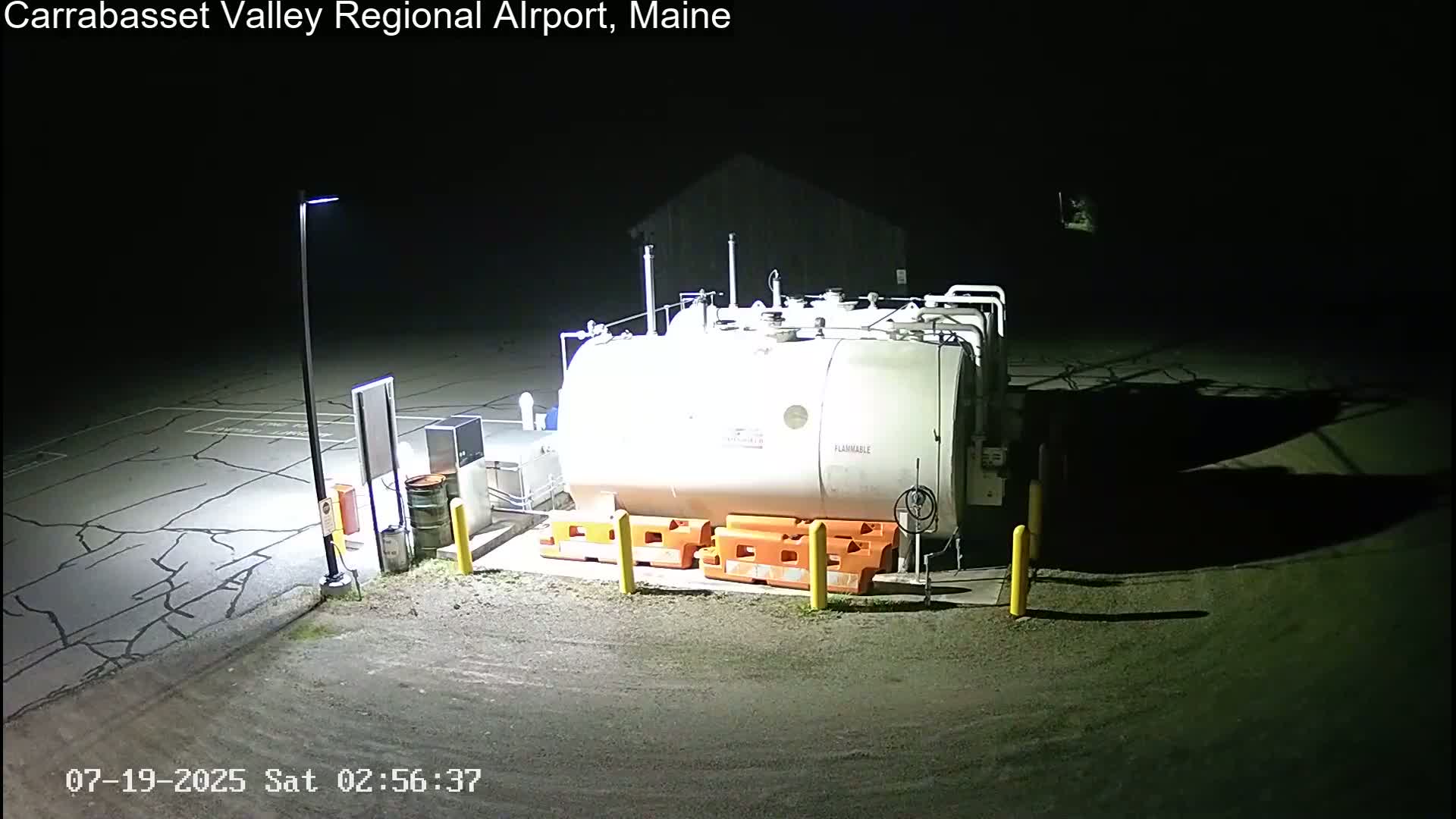 At night, a large white fuel tank sits in a mostly empty, cracked asphalt lot, illuminated by a single lamppost.