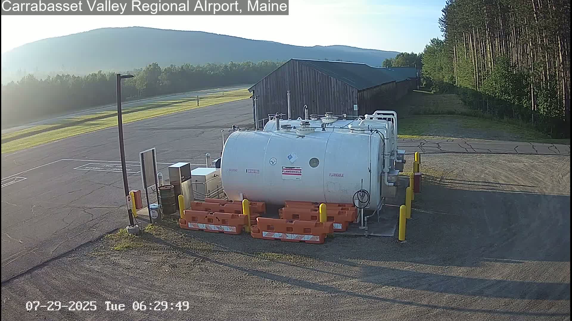 A large white cylindrical fuel tank sits in a paved area near a wooden building and a forested hillside under a clear sky.