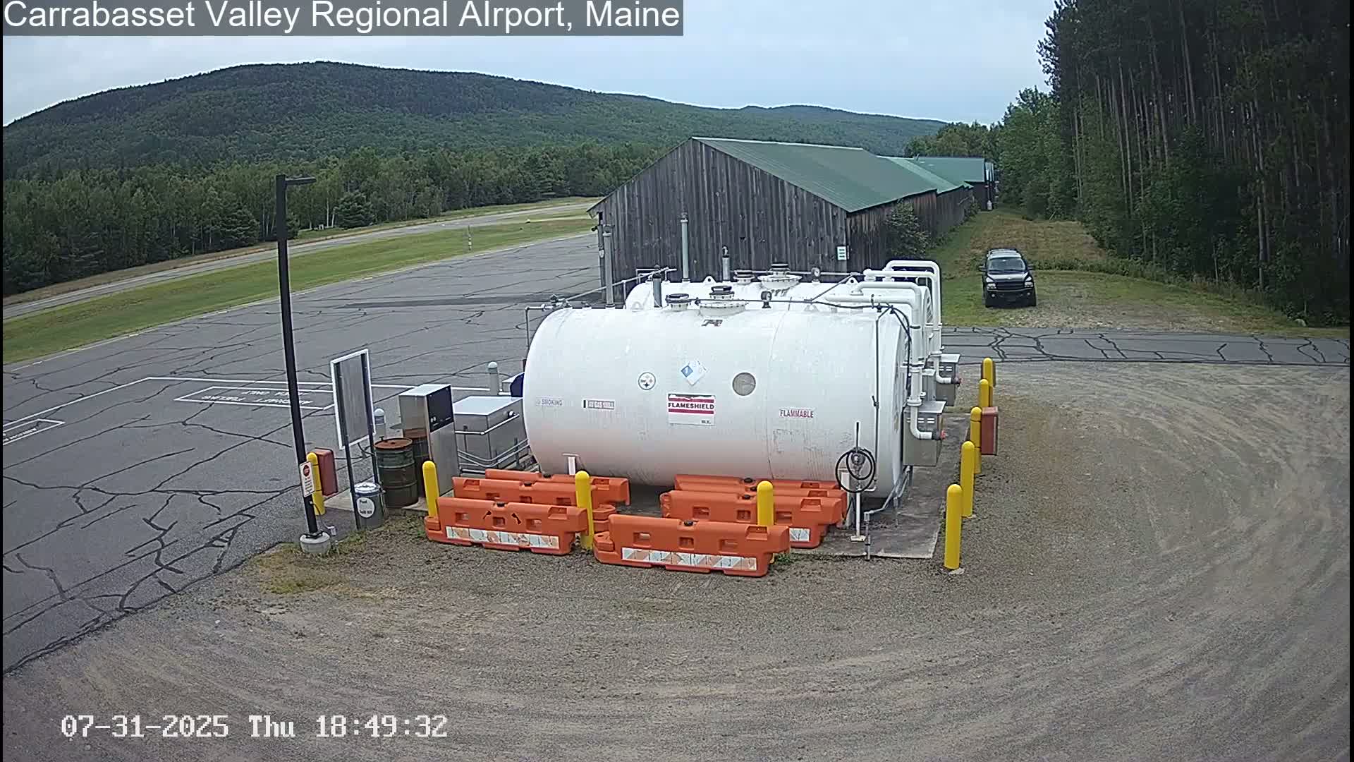 A large white fuel tank sits in a gravel parking lot near a dark-colored SUV, with a wood building and green forested hills in the background under an overcast sky.