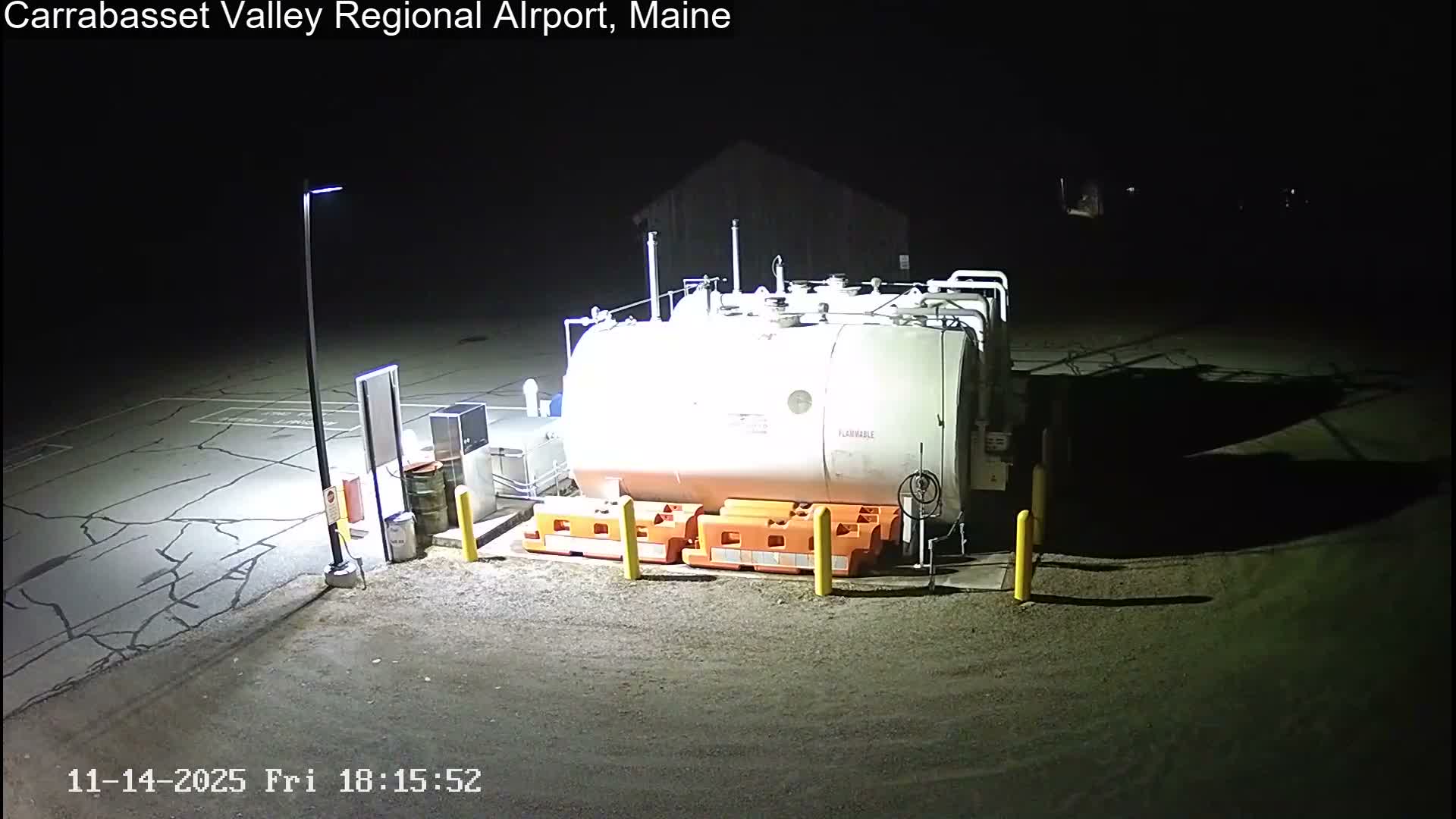 A large white fuel tank marked "FLAMMABLE" with orange safety barriers is illuminated by a street light on a cracked asphalt surface at night, under clear dark skies.