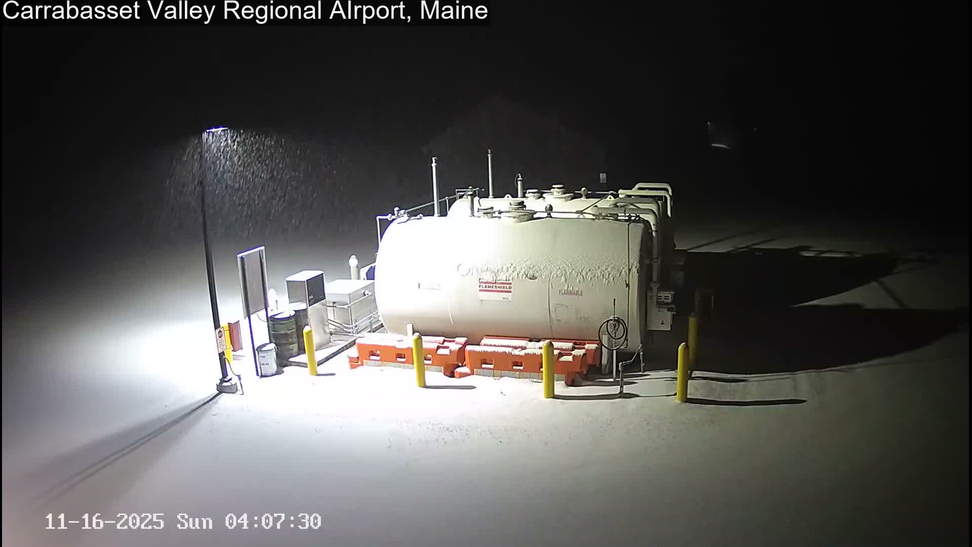 A large white fuel tank and surrounding equipment, partially covered in snow, are illuminated by a bright lamp post at night while snow falls.