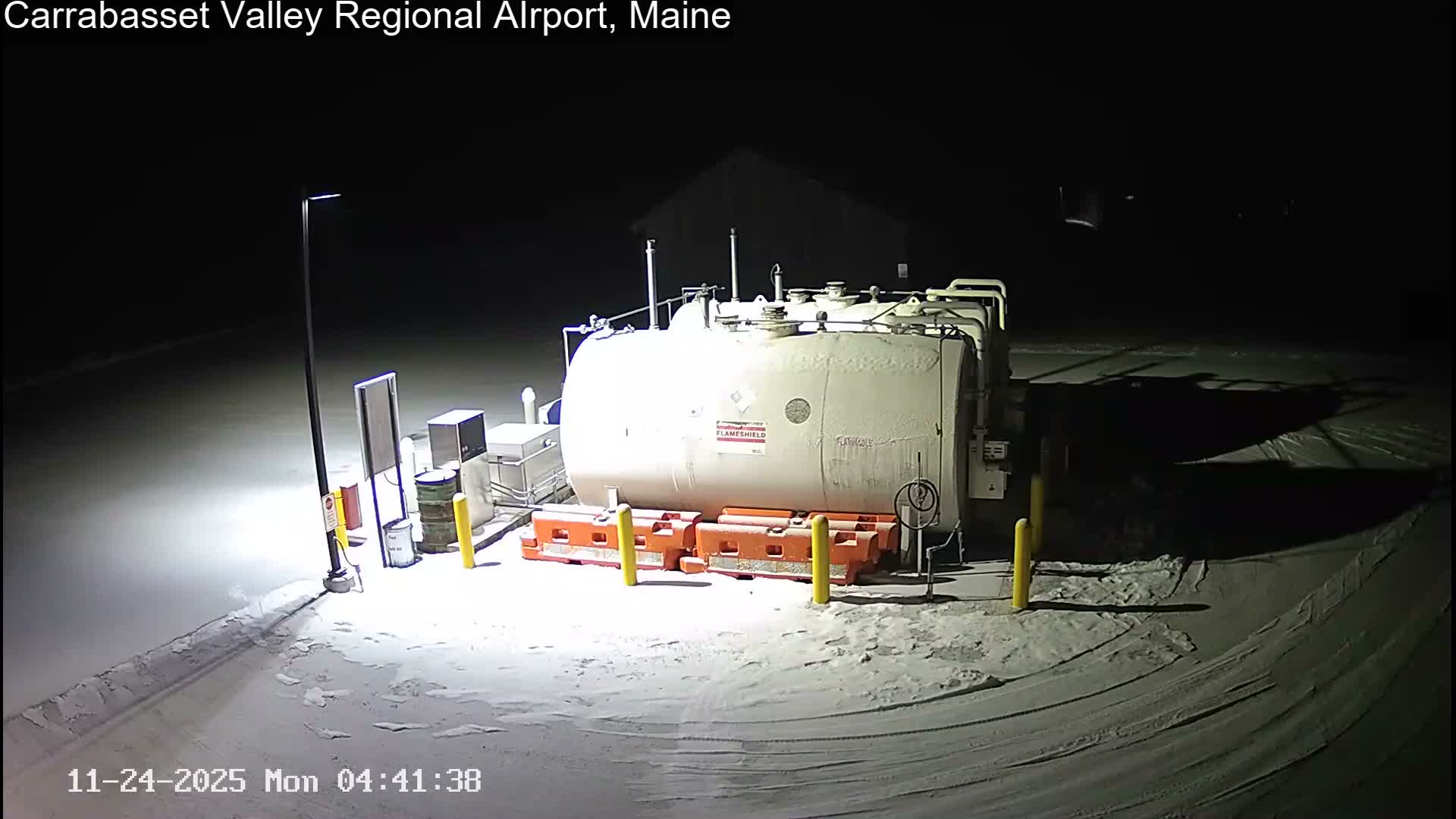 A light-colored fuel tank and associated equipment, including orange barriers and a lamppost, stand on a snow-covered ground under a dark night sky.