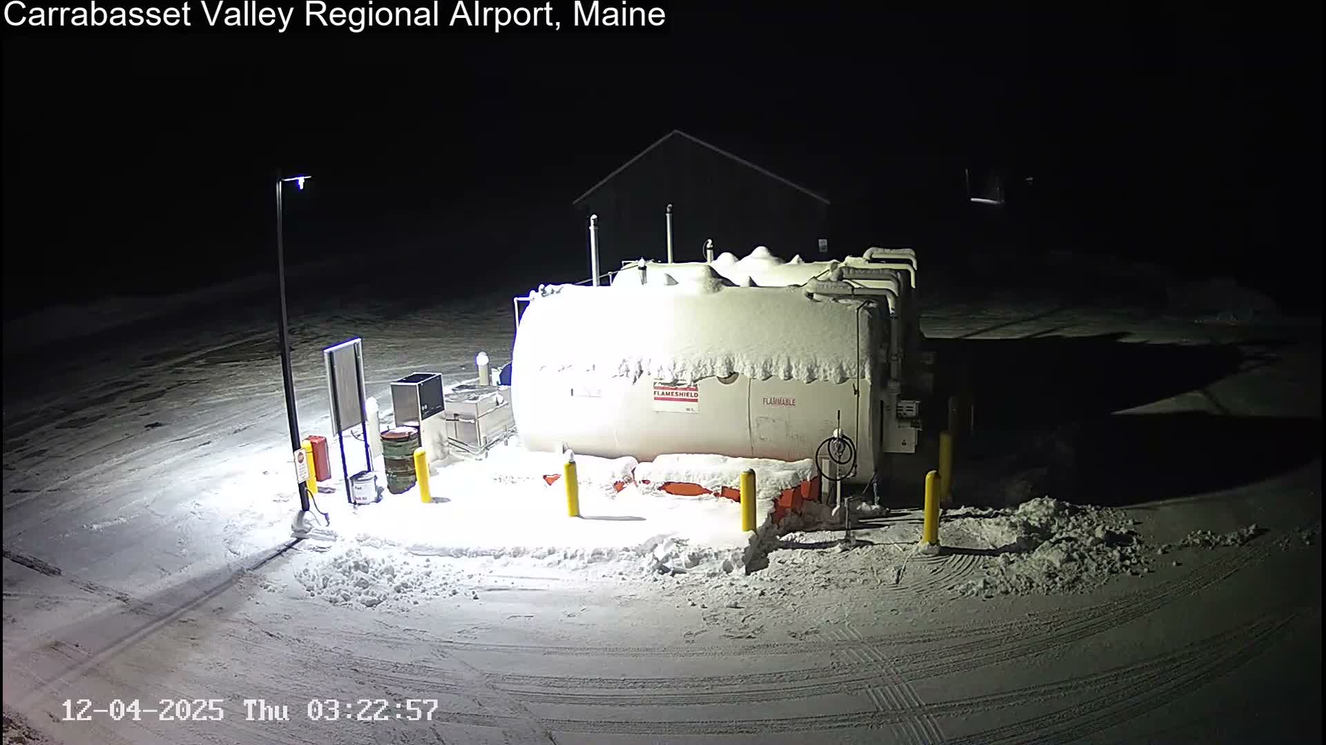 A lamp post illuminates a large, snow-covered white tank and its surrounding equipment at night, with the ground entirely blanketed in snow and marked by vehicle tracks.