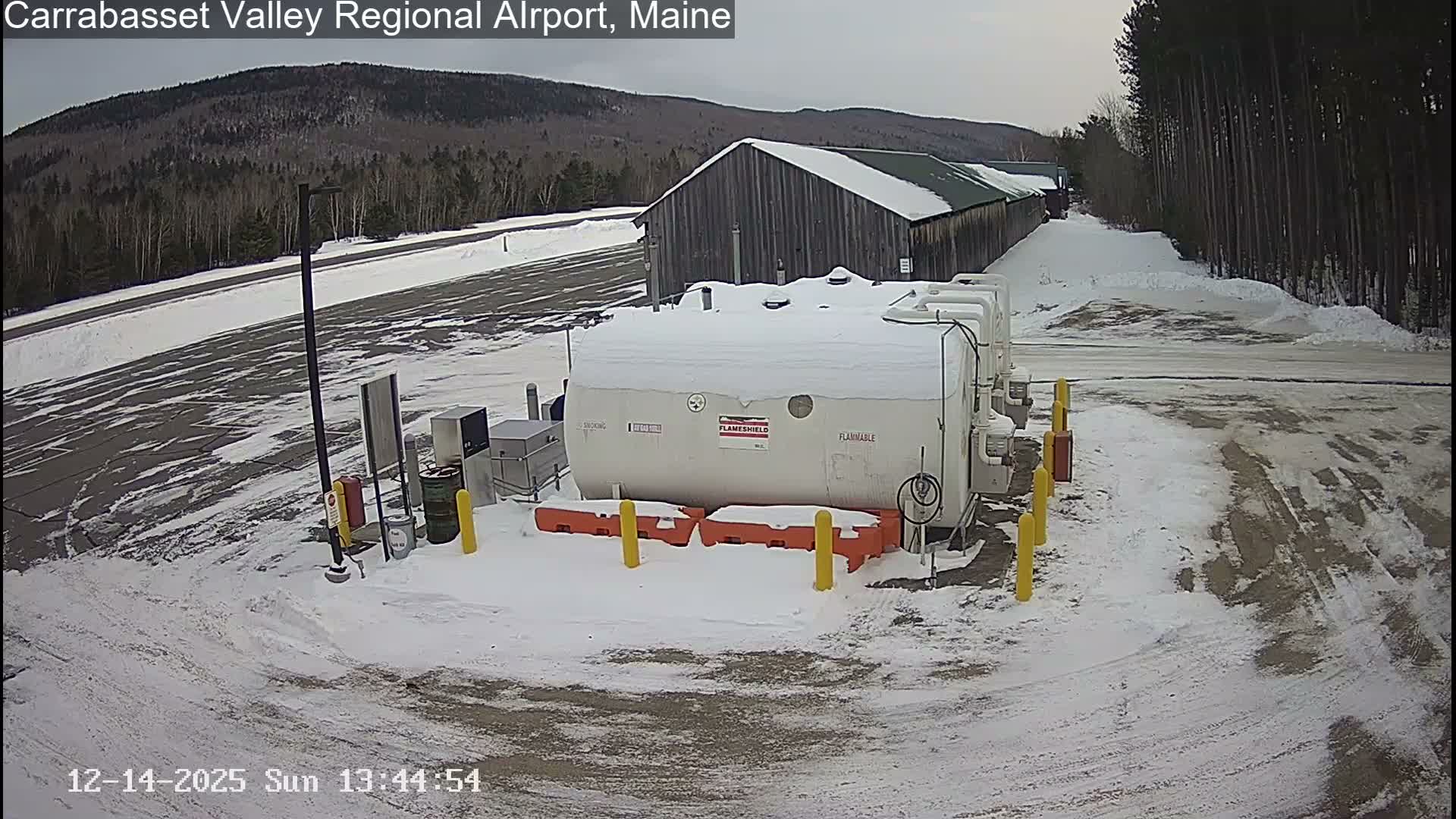 A lamp post illuminates a large, snow-covered white tank and its surrounding equipment at night, with the ground entirely blanketed in snow and marked by vehicle tracks.