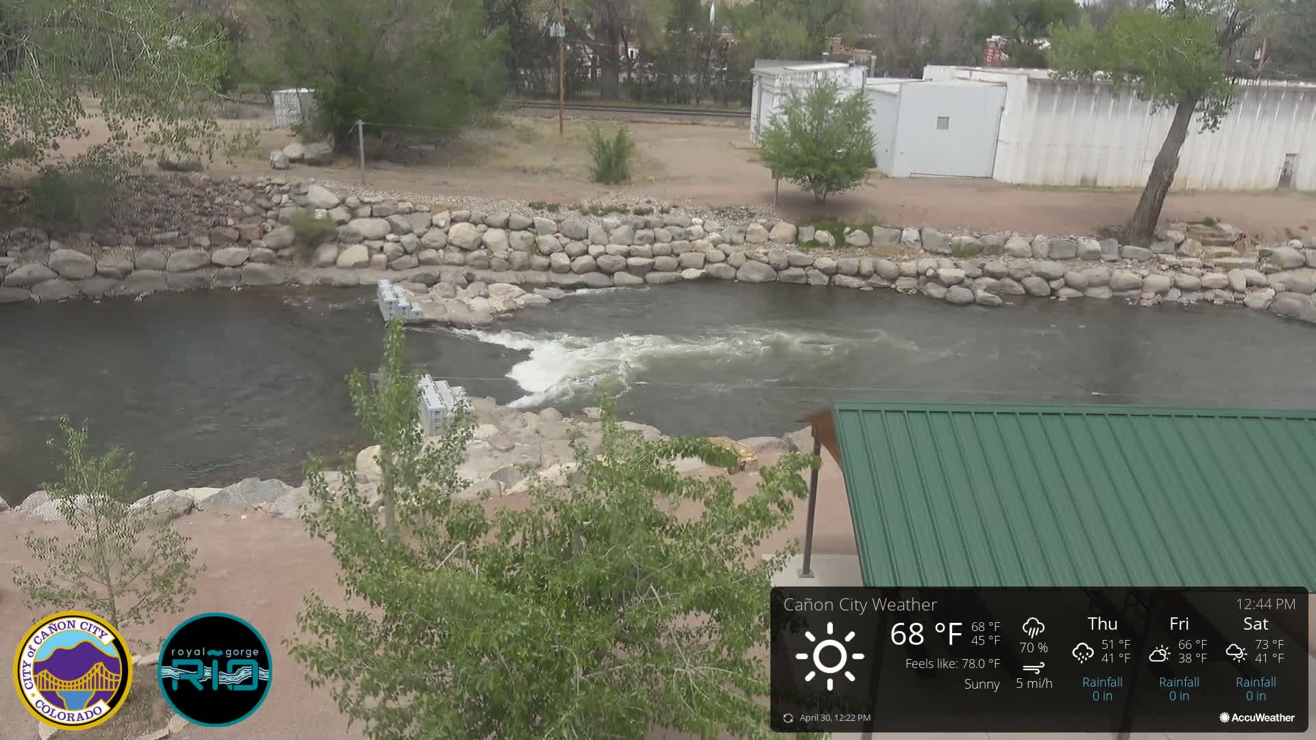 A sunny day reveals a river flowing over a low rock dam, partially obscured by vegetation and a green metal roof.