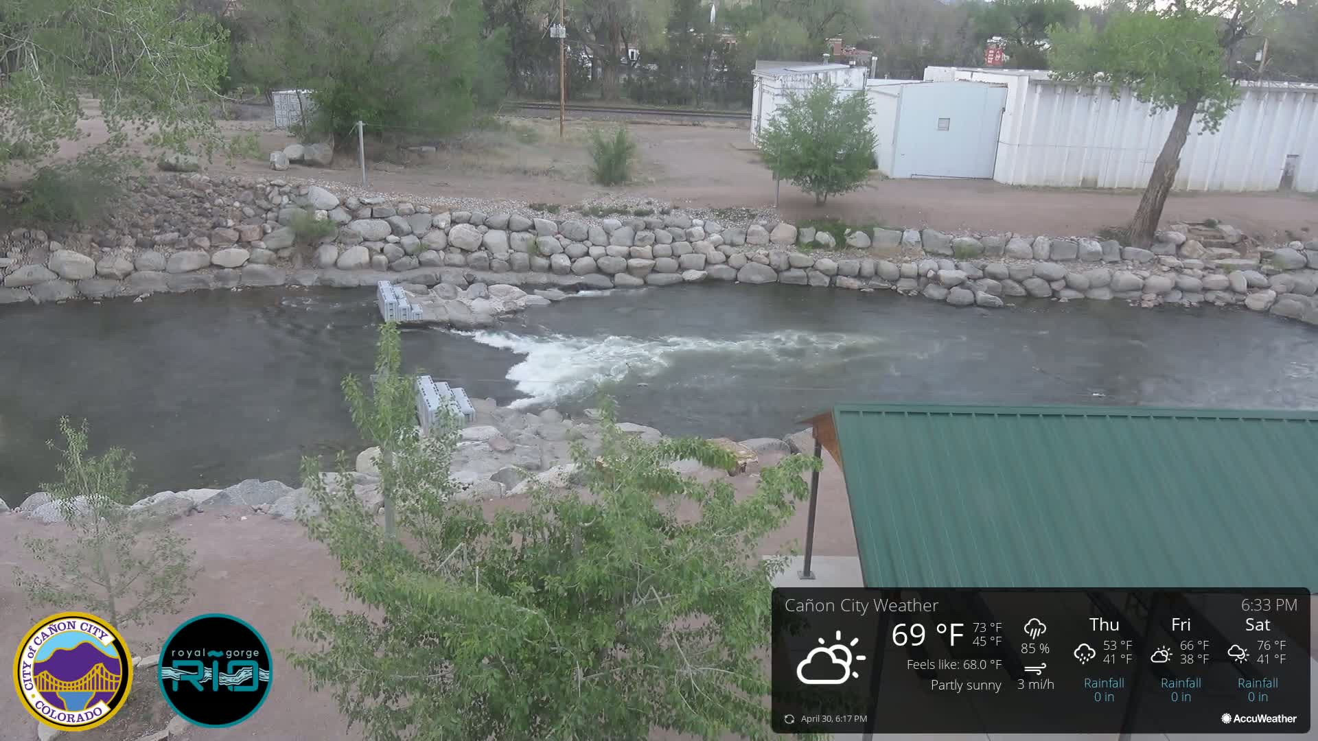 A partly sunny view of a river flowing past a rock wall and a green-roofed structure.