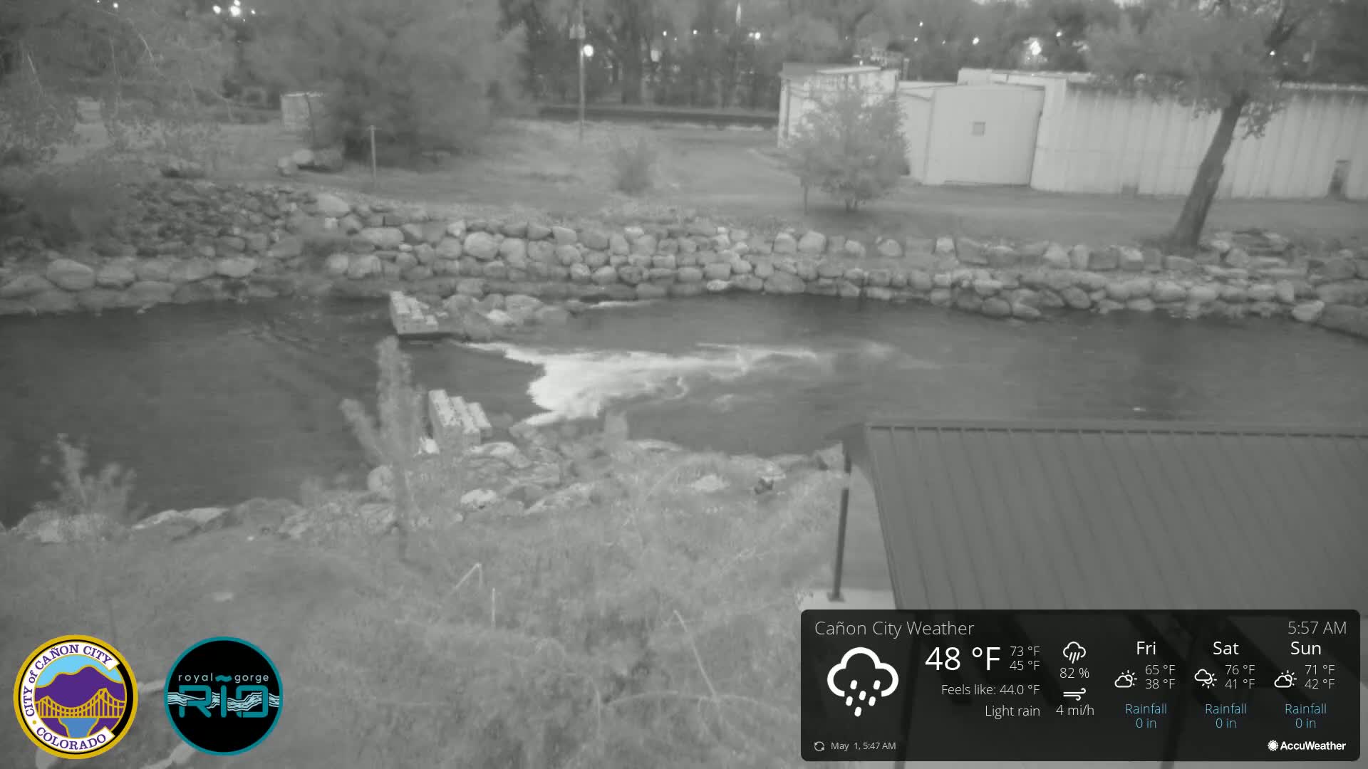 A nighttime, grayscale image shows a rocky riverbank with a flowing river under light rain, alongside a building and some vegetation.