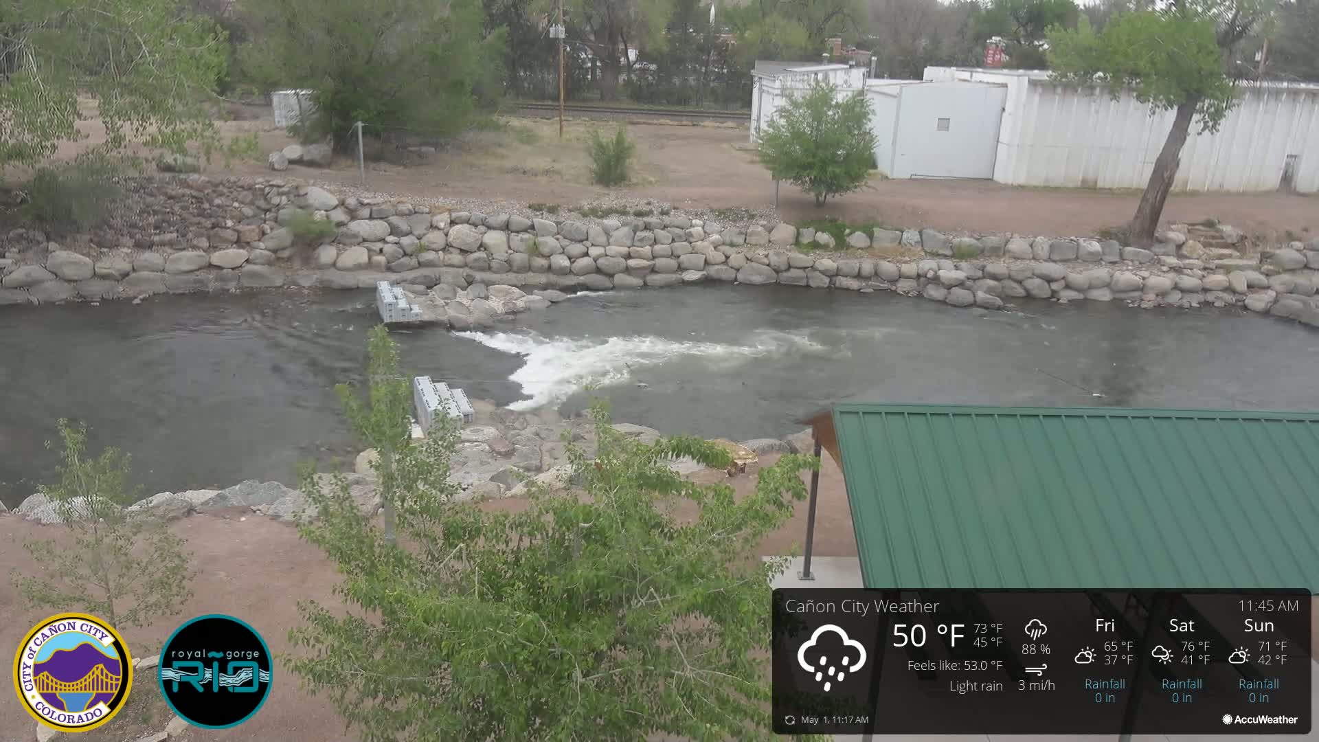 A river flows through a rocky area with light rain, under a partly cloudy sky; a portion of a green roofed structure is visible in the lower right.