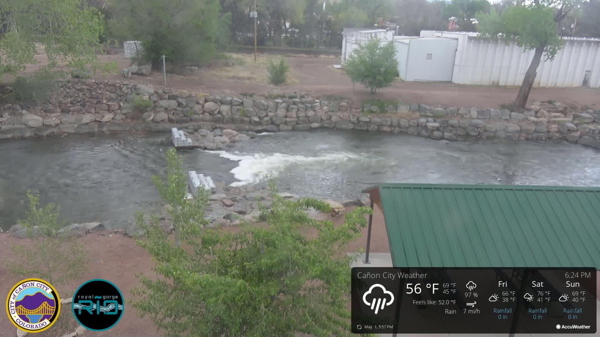 A river flows past a rock wall and under a green roofed structure; it's raining.
