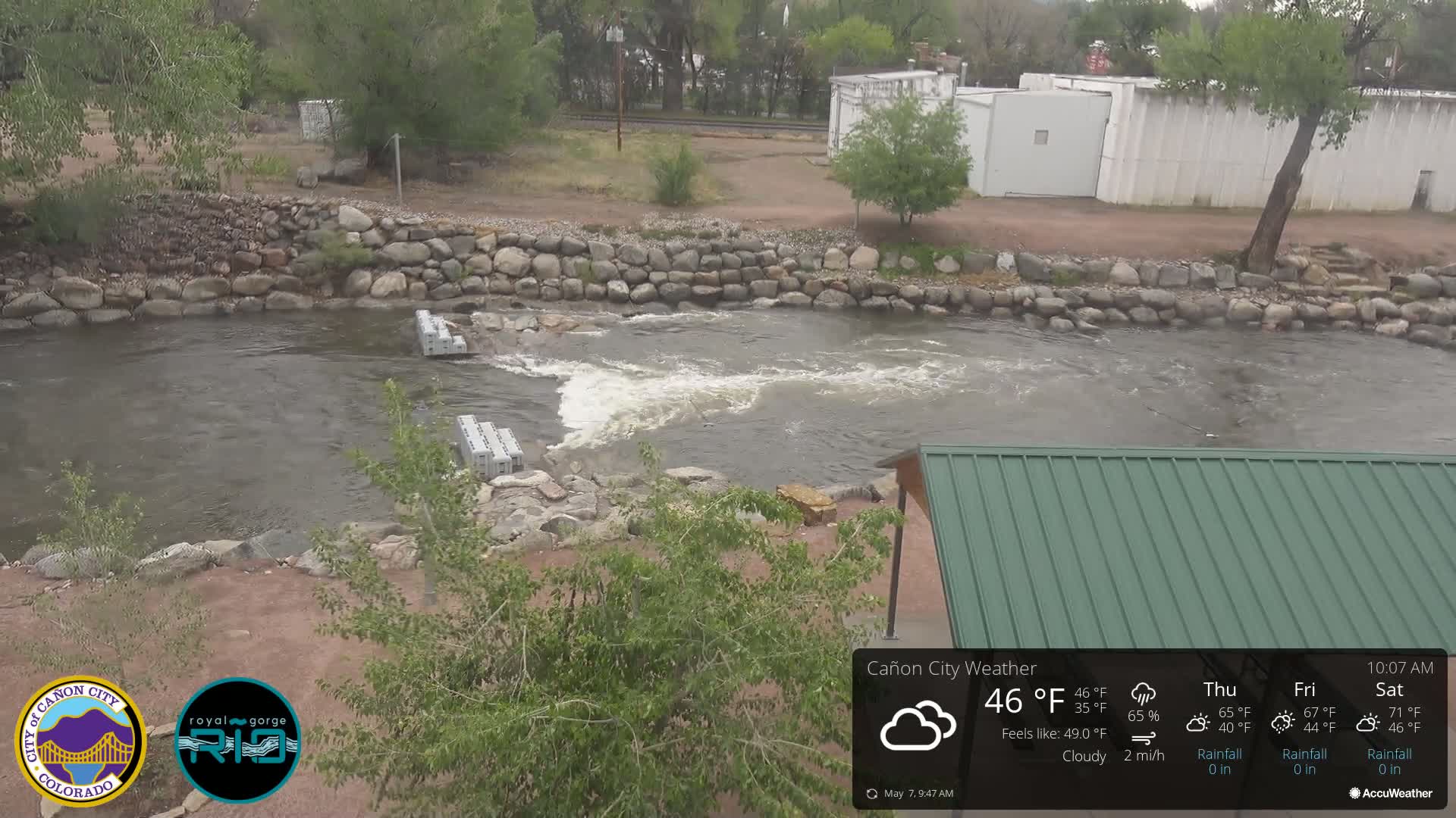 A cloudy day shows a rocky riverbank with a section of calm water and a small, rushing area where several gray rectangular structures are partially submerged; a green metal roofed structure is visible in the lower right.