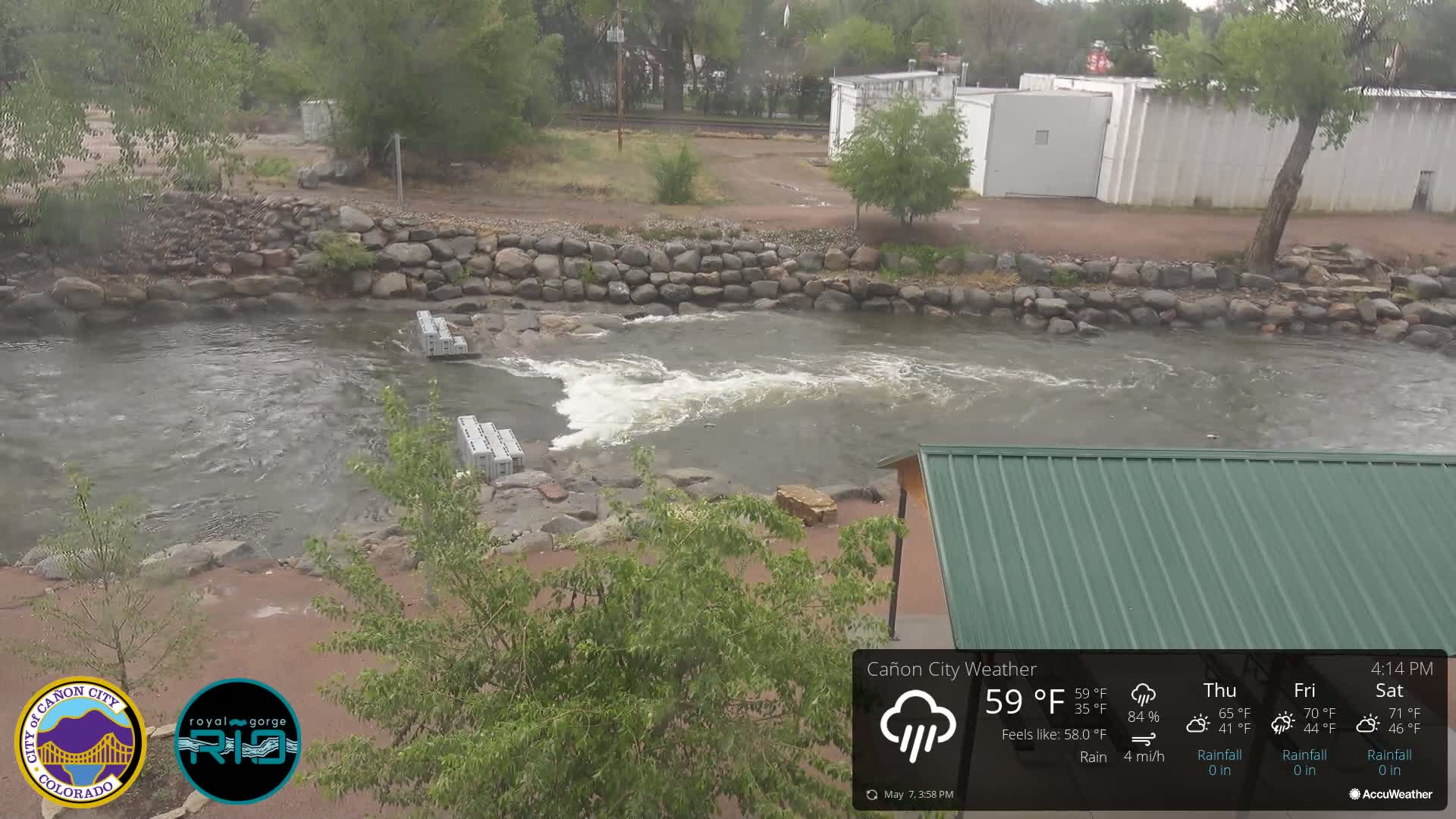 A rocky river flows under a partly cloudy sky during a light rain, with several concrete blocks partially submerged near the riverbank.