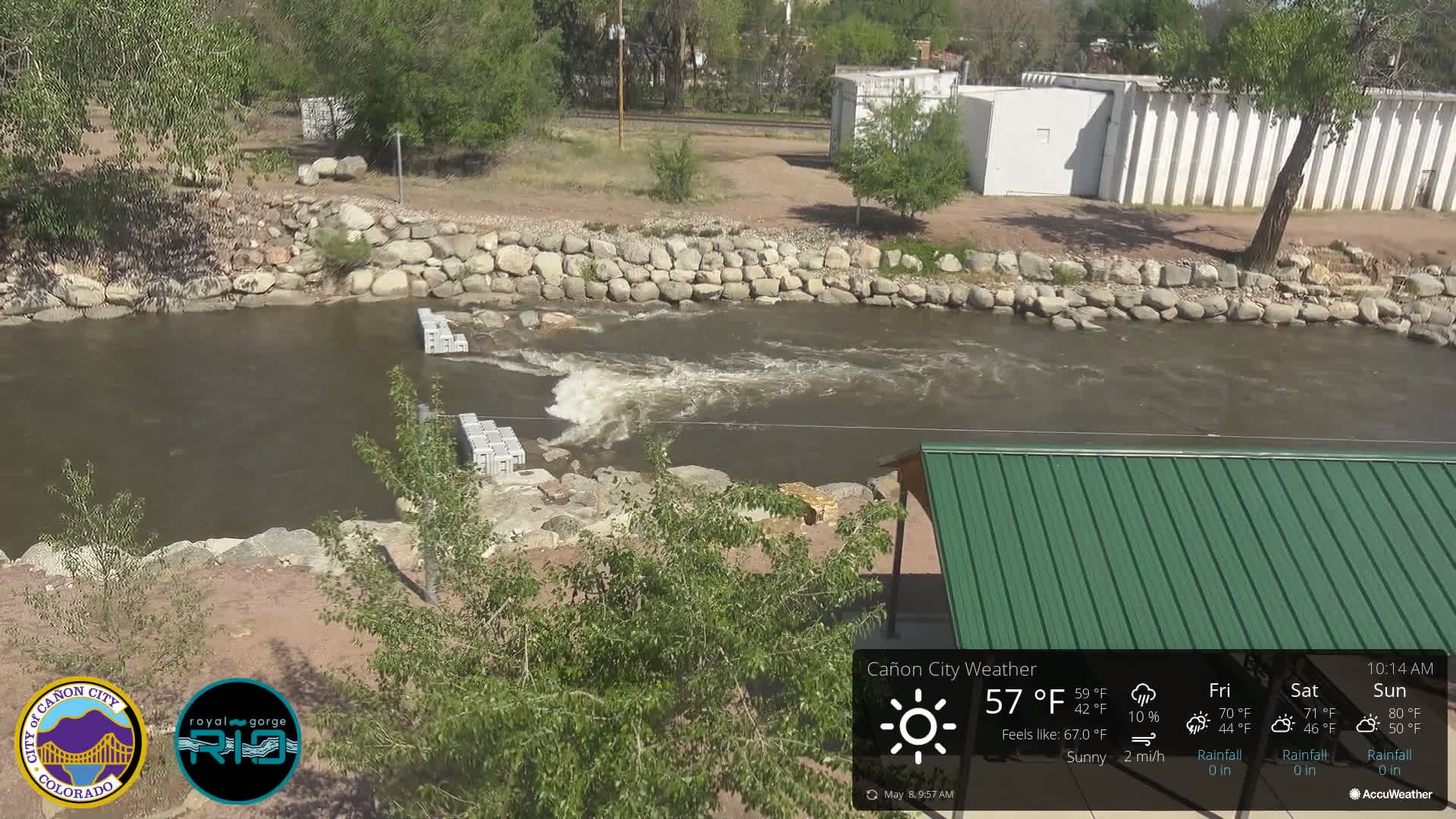 A sunny day shows a river flowing around gray concrete blocks, bordered by rocks and green vegetation, with a green roofed structure in the lower right.